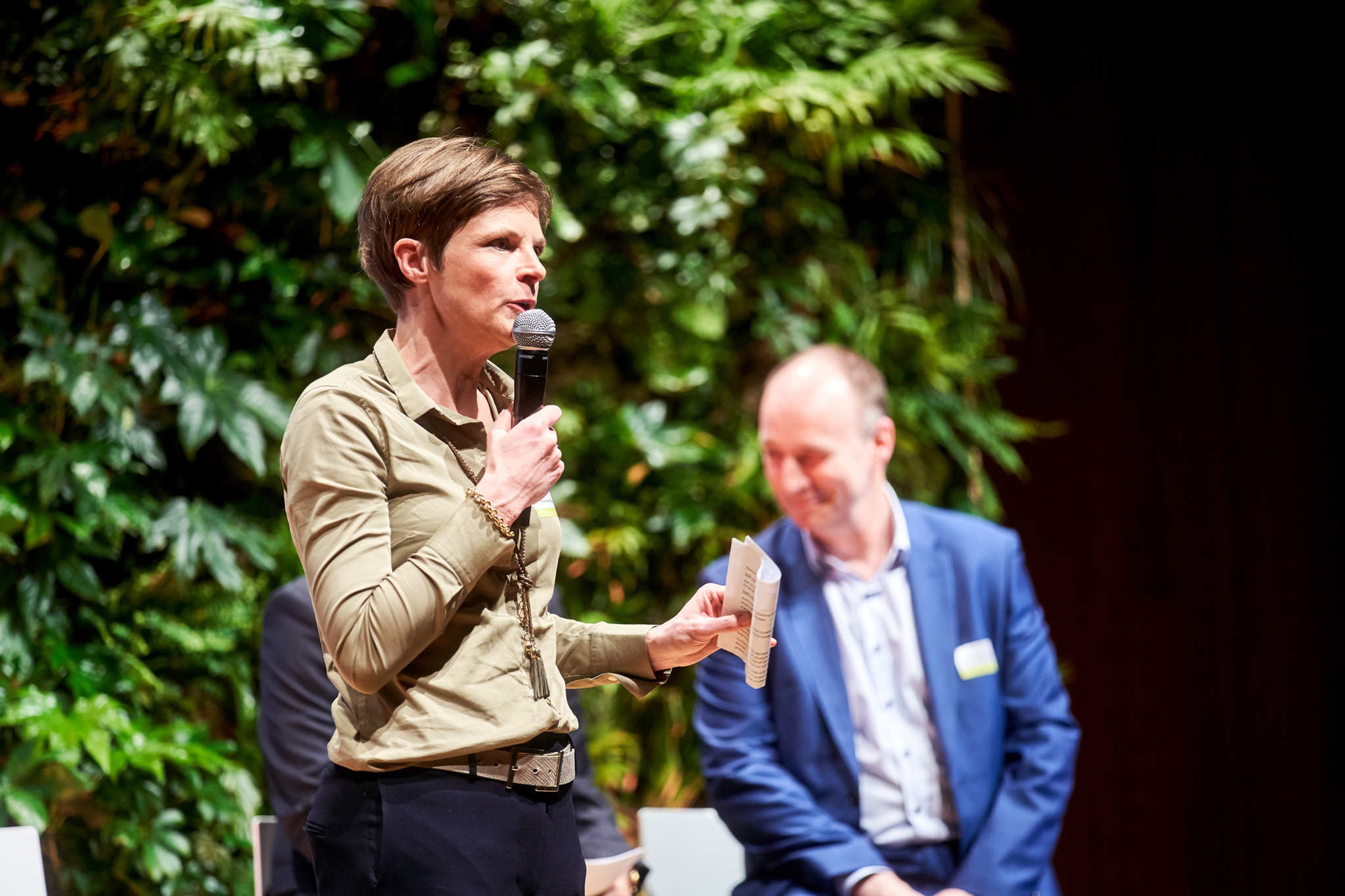 Een vrouw spreekt in een microfoon op het podium, terwijl ze een papier vasthoudt, met een weelderige groene plantenwand achter haar. Een man in een blauw pak luistert aandachtig.