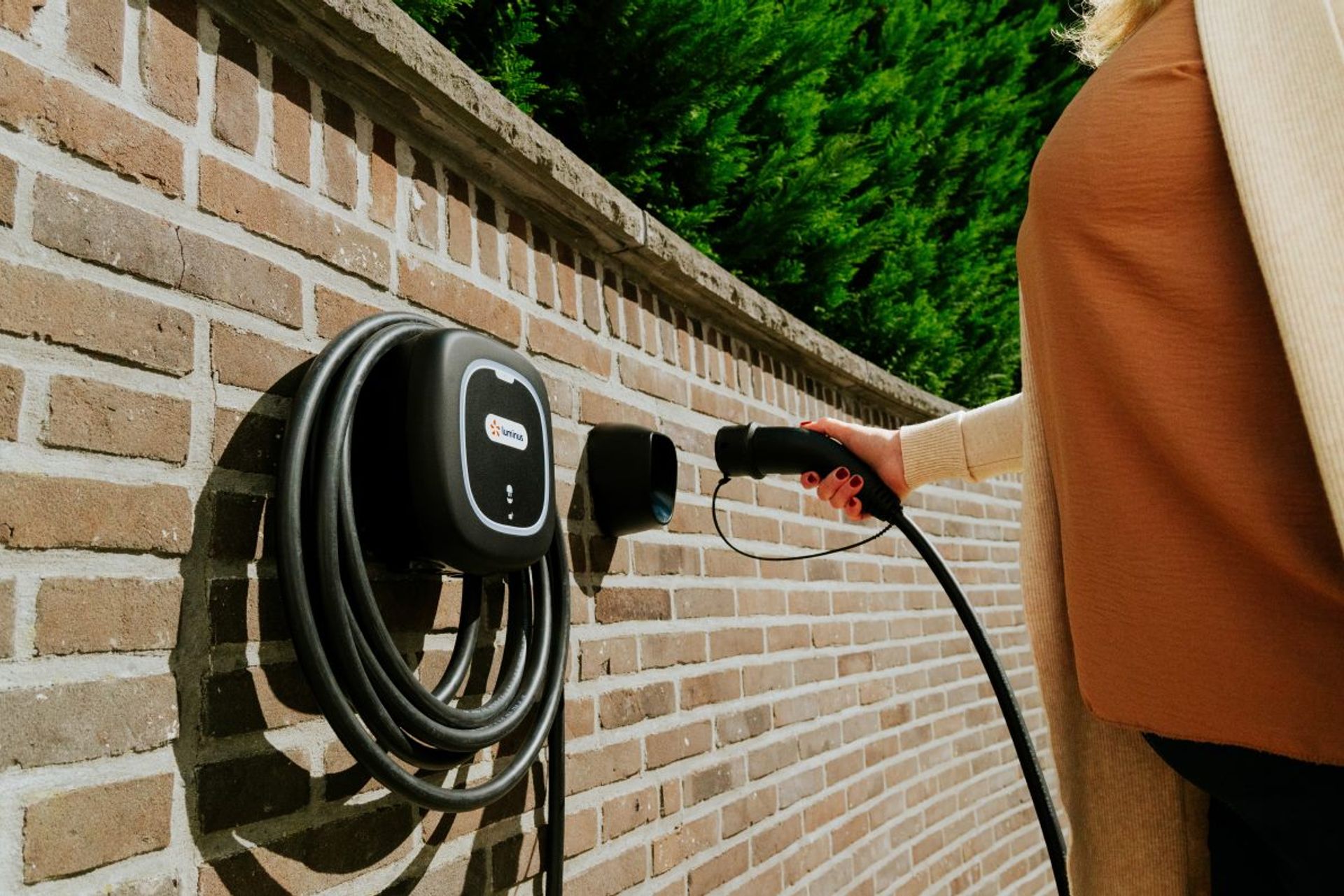 Bakstenen muur met wallpoint: stopcontact en laadkabel voor elektrische wagen, vrouw met oranje top steekt de stekker in stopcontact