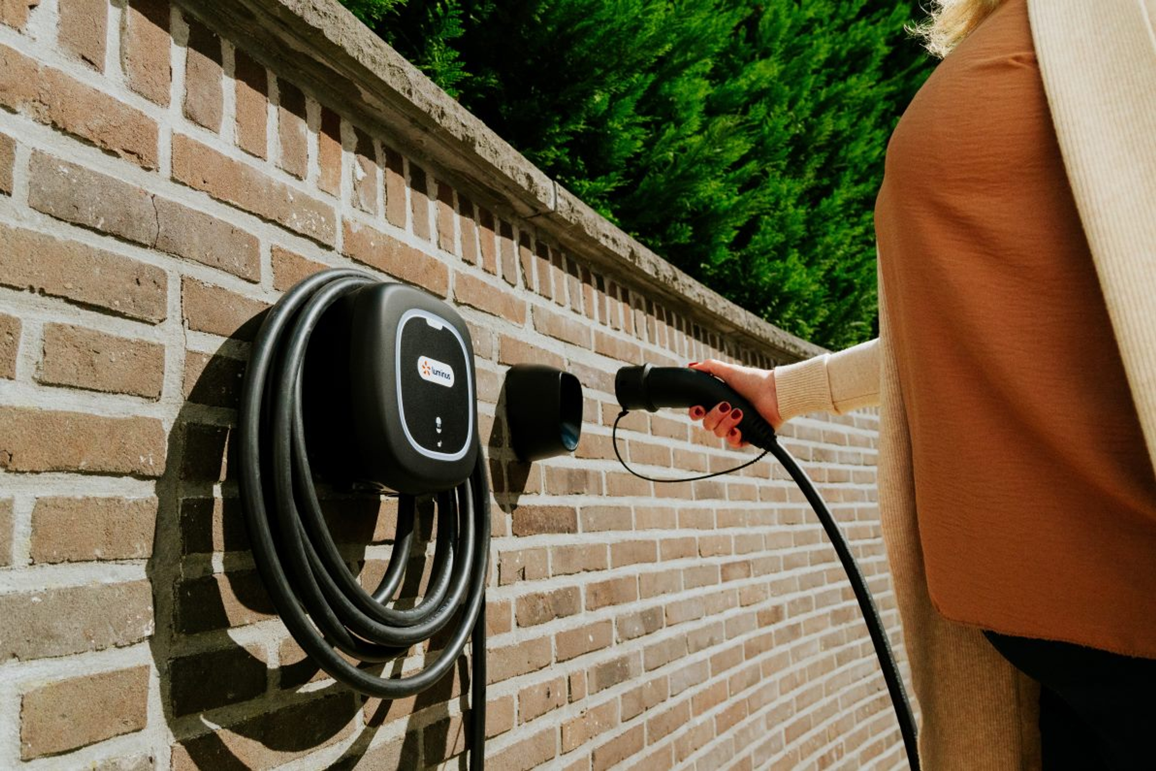 Bakstenen muur met daarop een zwarte wallbox laadpunt. Dame met bruine top steekt de stekker in.