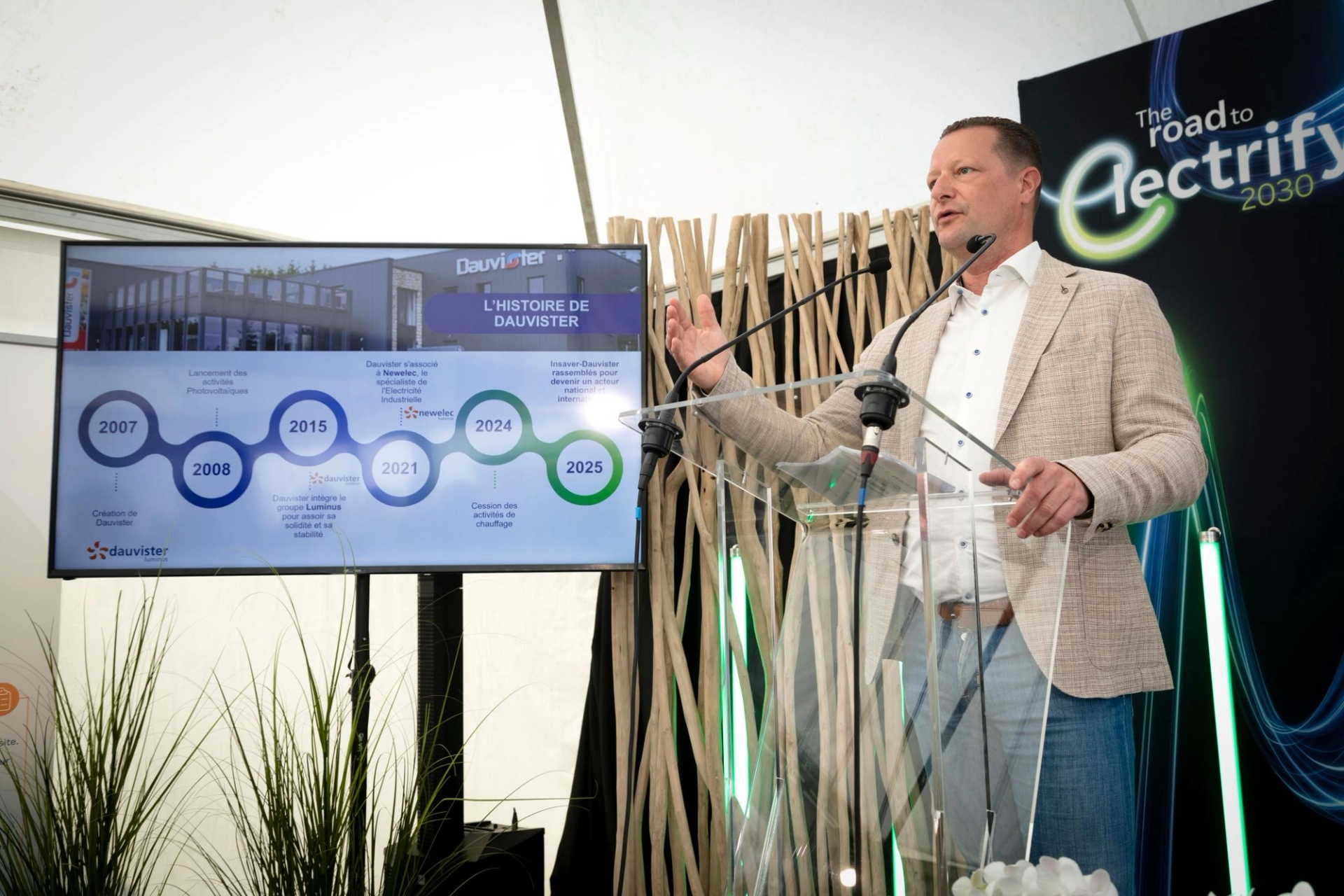 Un homme en blazer beige s’exprime depuis une tribune, à côté d’un écran sur lequel une frise chronologique est affichée.