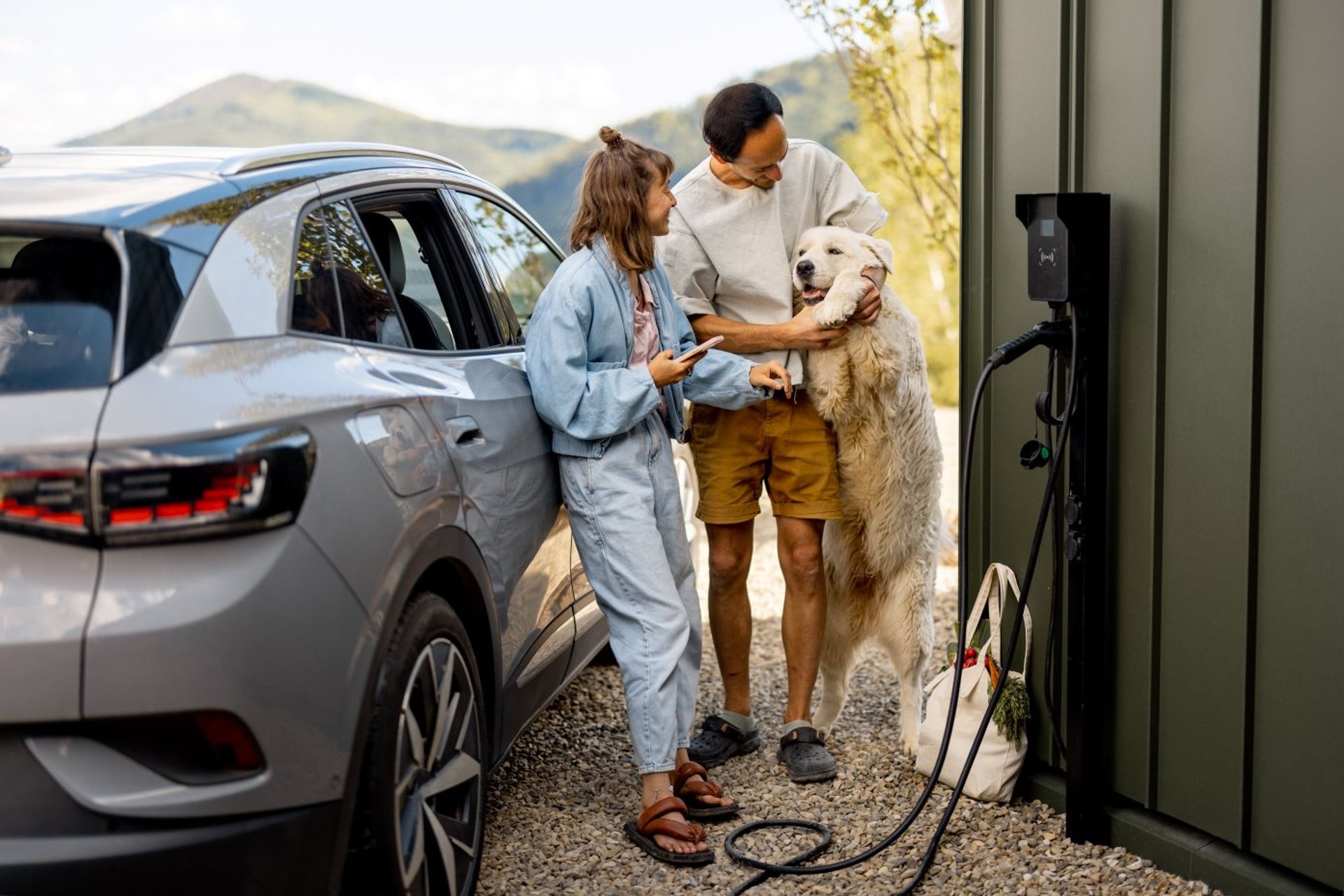 Elektrische wagen bij een oplaadpunt aan zwarte muur. Jong koppel met bleke grote hond staat erbij.