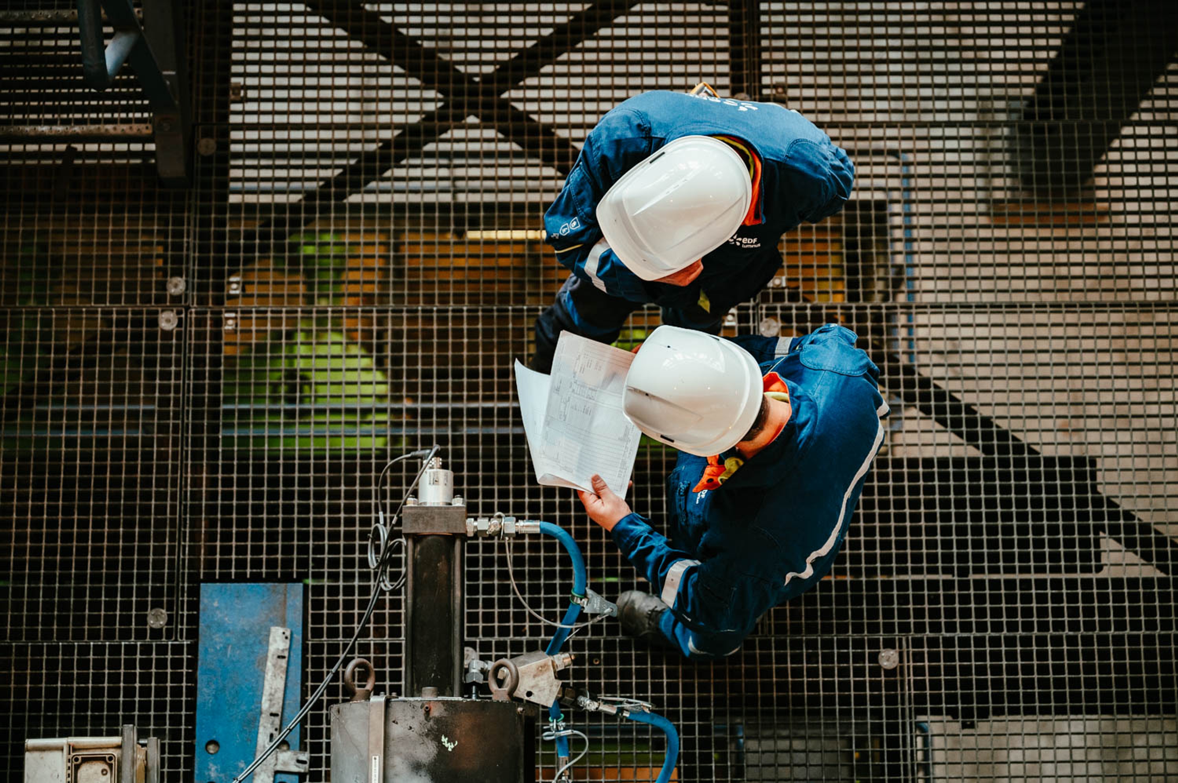 Twee technici met witte helm staan op roostervloer en kijken op papieren, bovenaanzicht