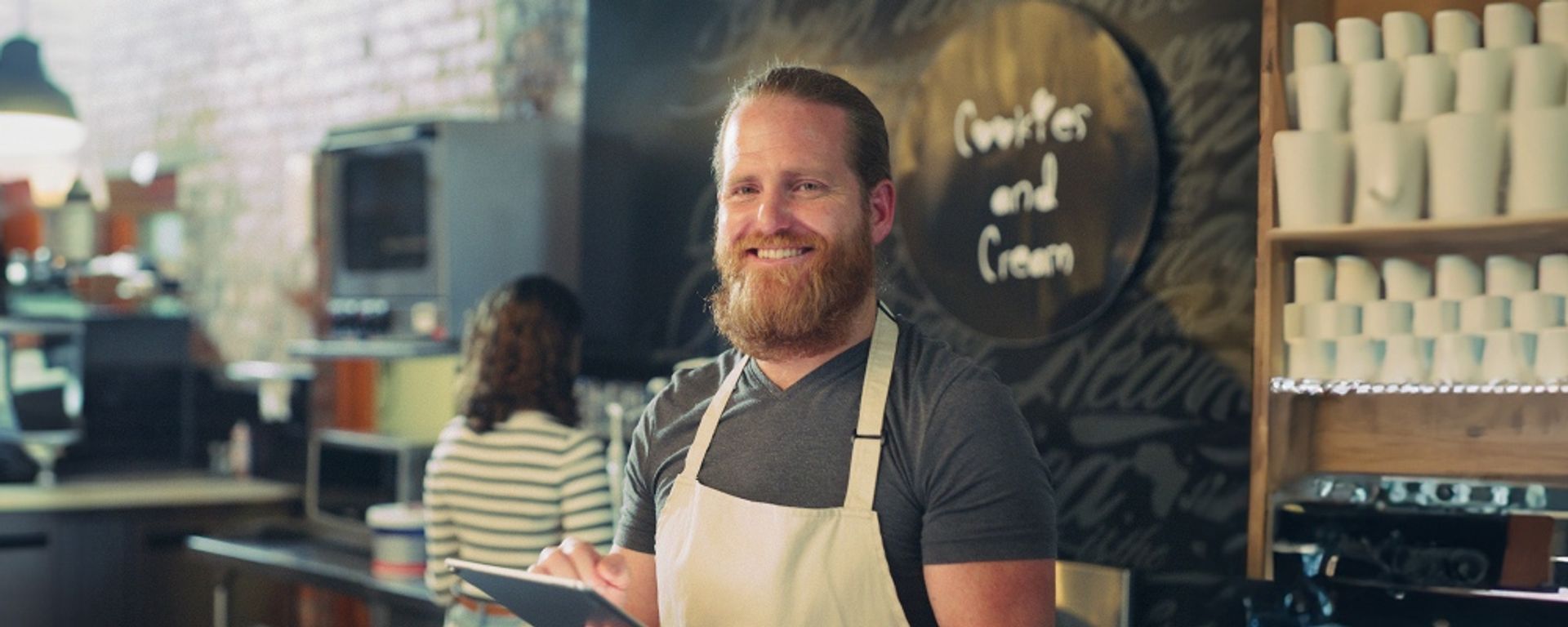 Man met baard en schort lachend achter de toog van een koffiebar, met tablet in de hand.