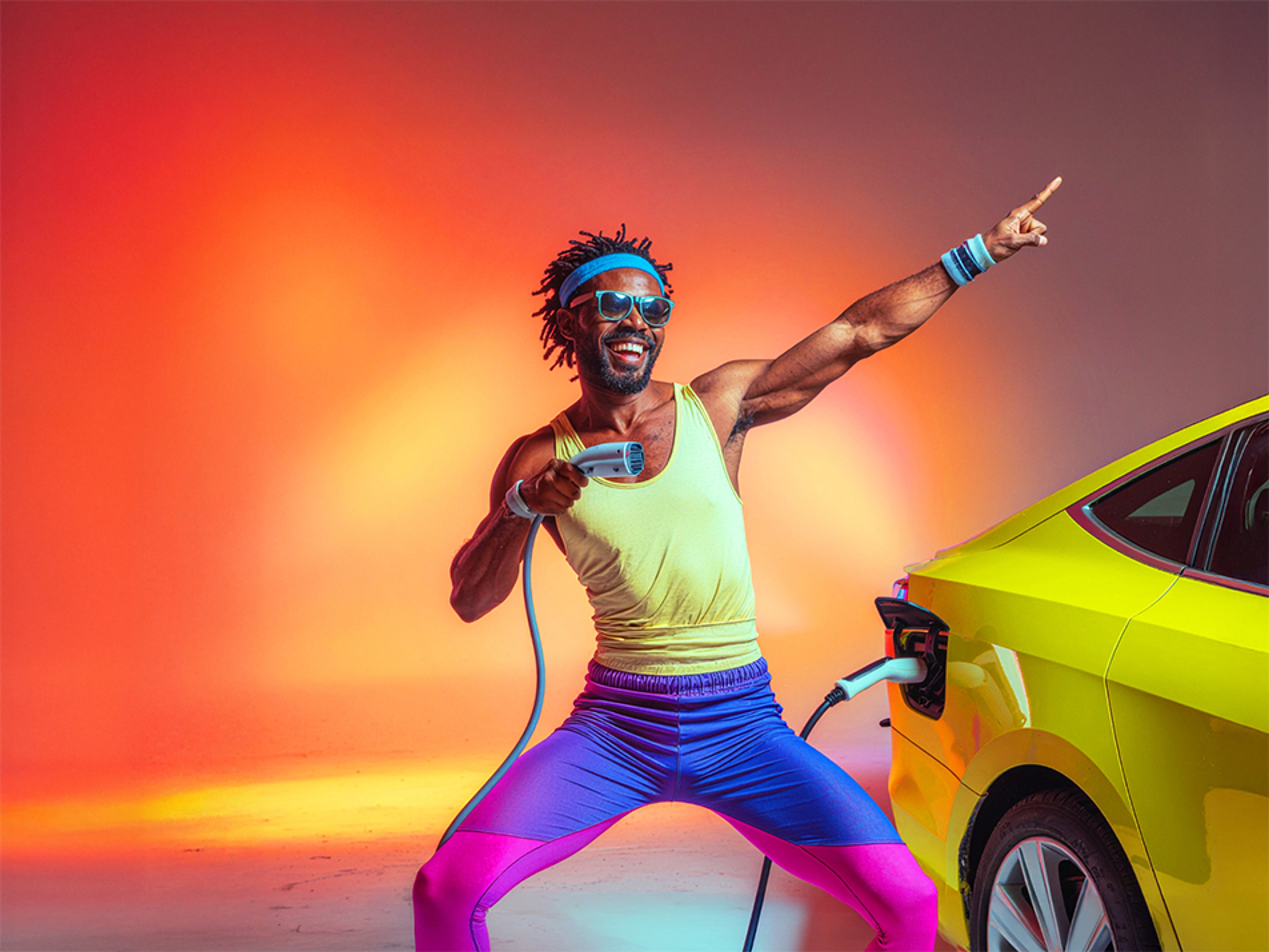 Dynamische kleurrijke man in geel shirt met blauw-roze broek staat lachend en zingend bij haar wagen die aan het opladen is. 