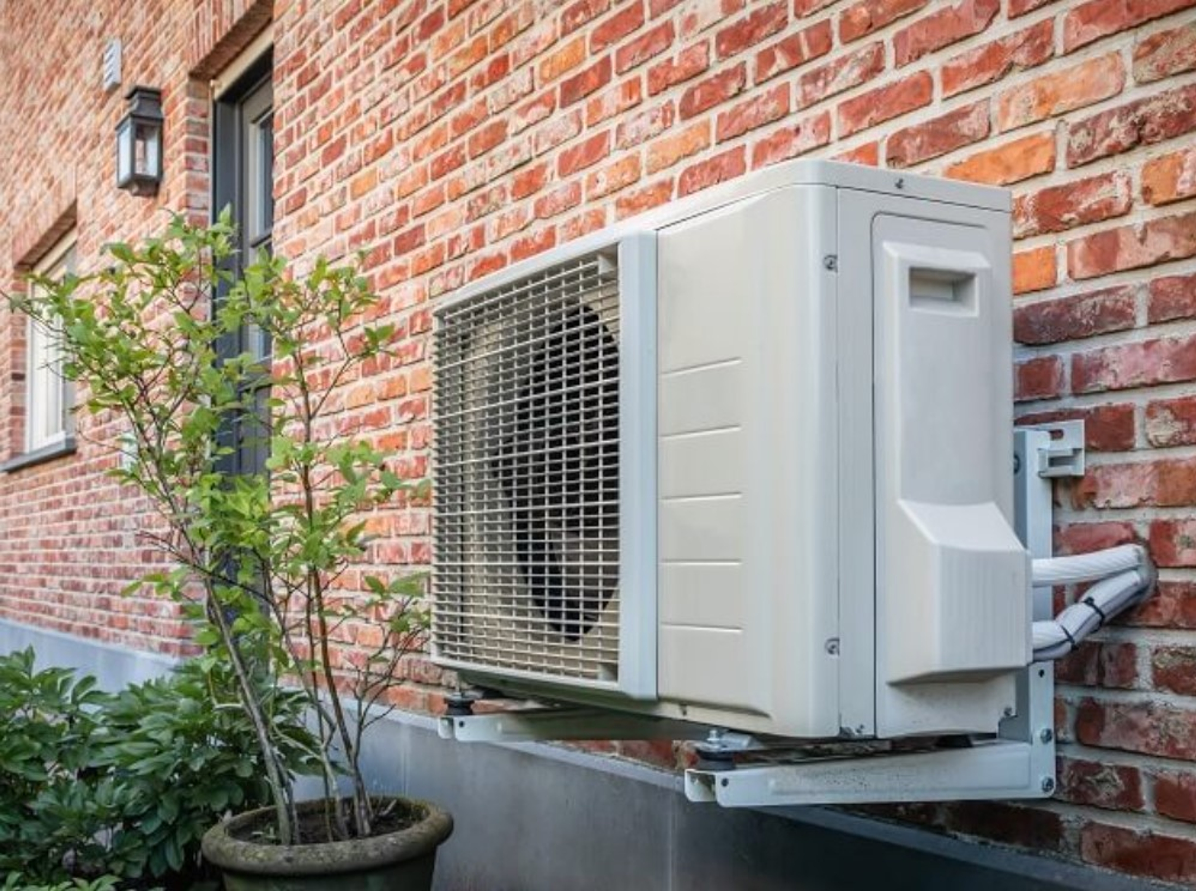 Pompe à chaleur air-eau fixée au mur en briques d’une maison, installée sur un support métallique, avec une plante en pot à proximité
