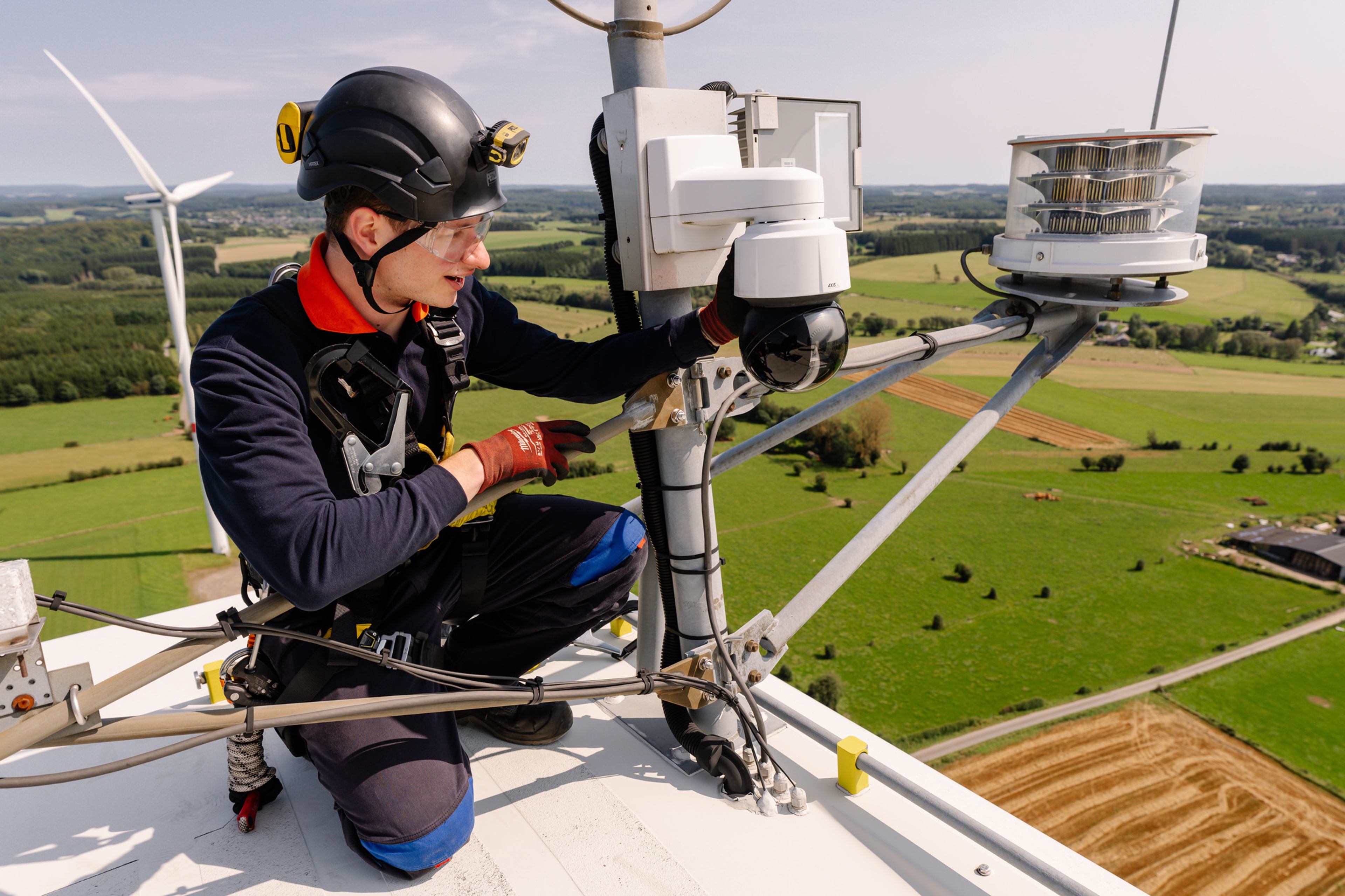 Un technicien équipé d'un équipement de sécurité travaille au sommet d'une éolienne, inspectant l'équipement avec en toile de fond des champs verdoyants et une autre éolienne au loin.