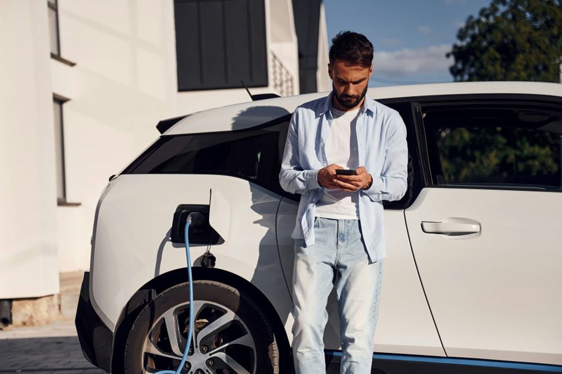 Man met baardje en bleke jeans staat bij zijn witte e-wagen, kijkt op zijn phone.