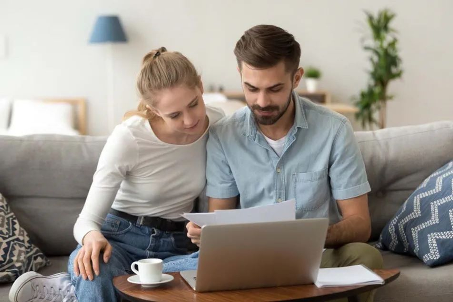 Couple assis sur un canapé, consultant ensemble des documents papier devant un ordinateur portable posé sur une table basse avec une tasse de café