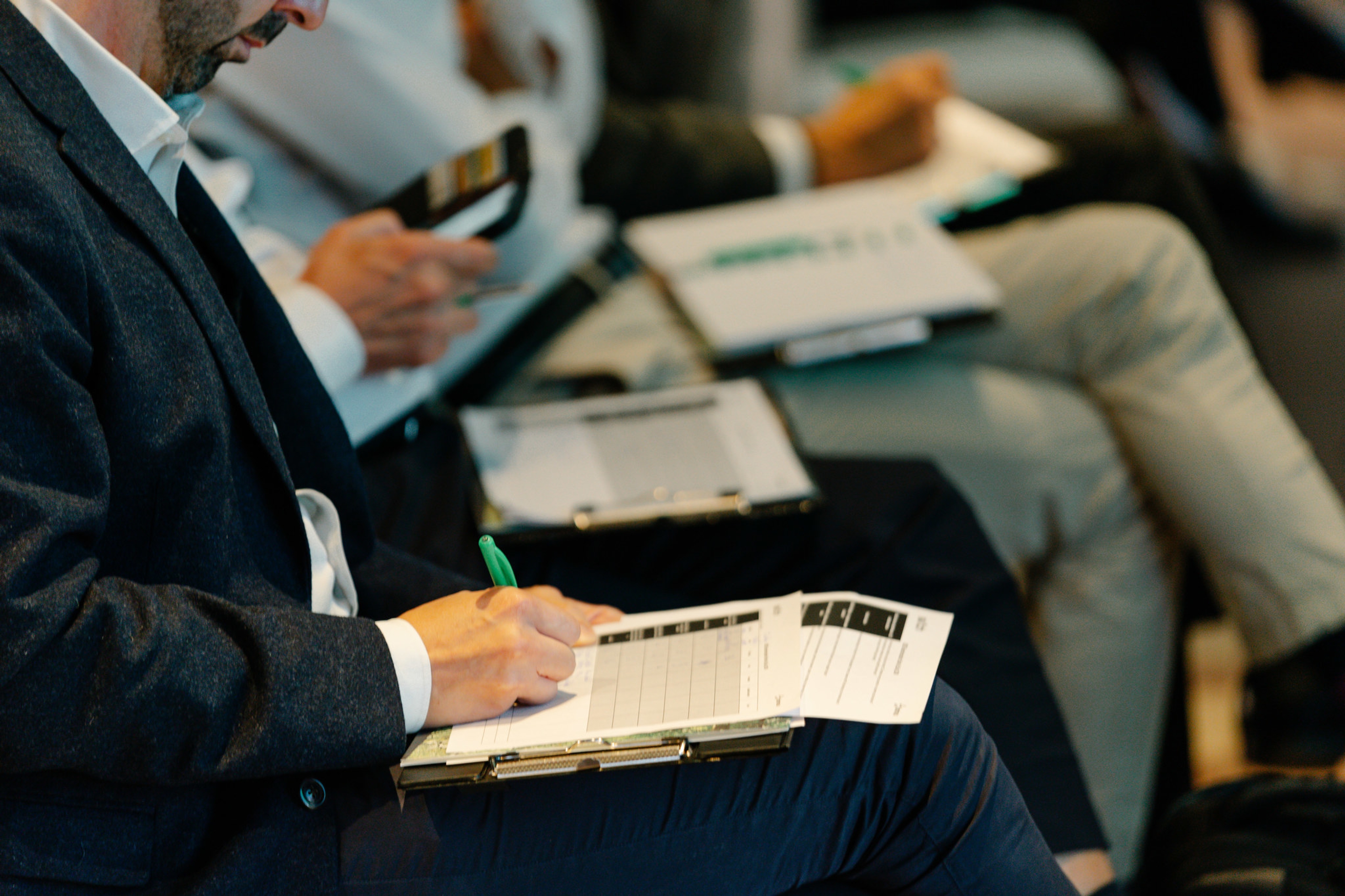 Gros plan sur des personnes assises prenant des notes sur des blocs-notes lors d'une conférence. Un homme écrit avec un stylo vert.