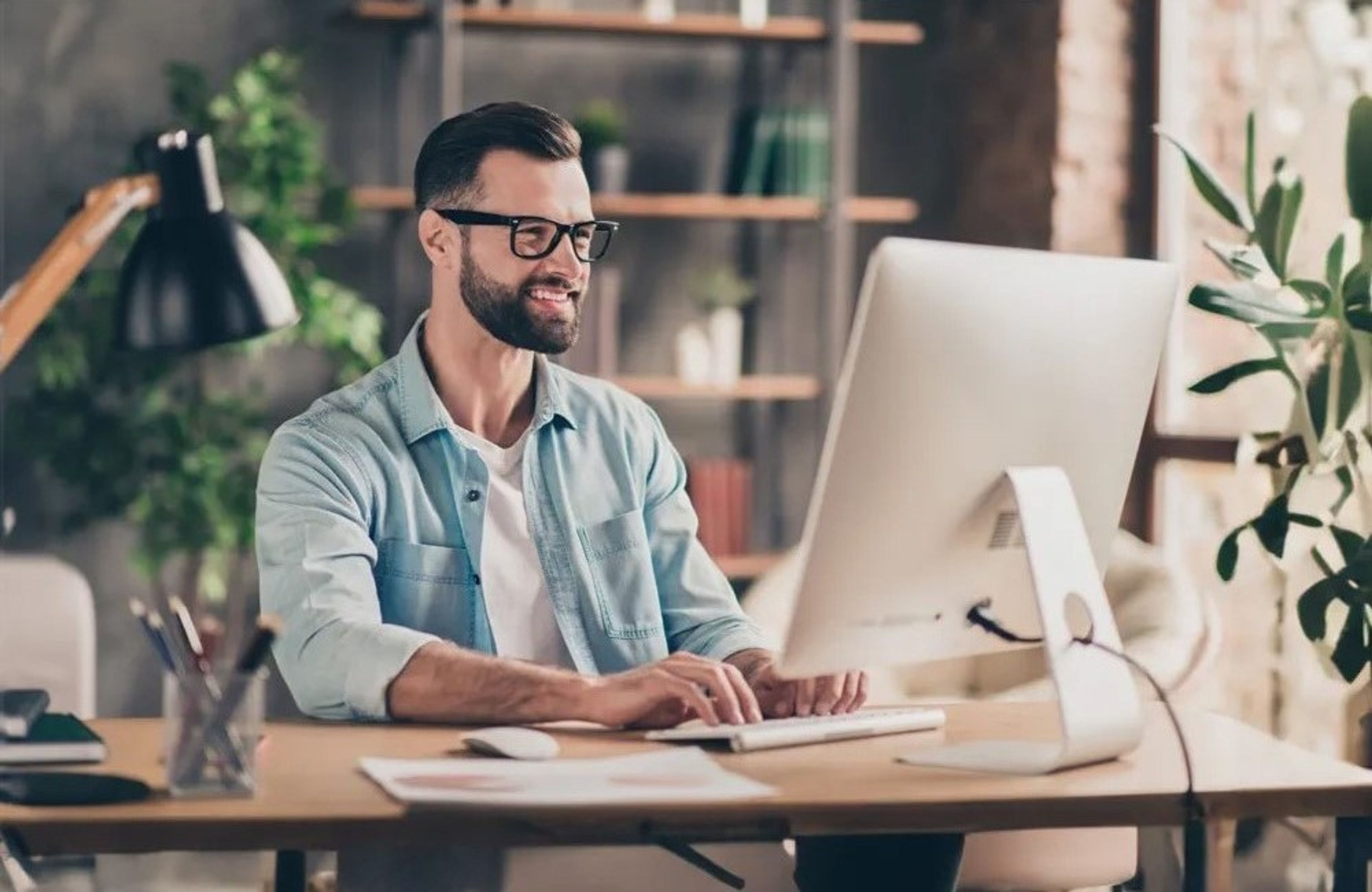 Jonge man met baard en jeanshemd zit lachend achter de computer te werken.