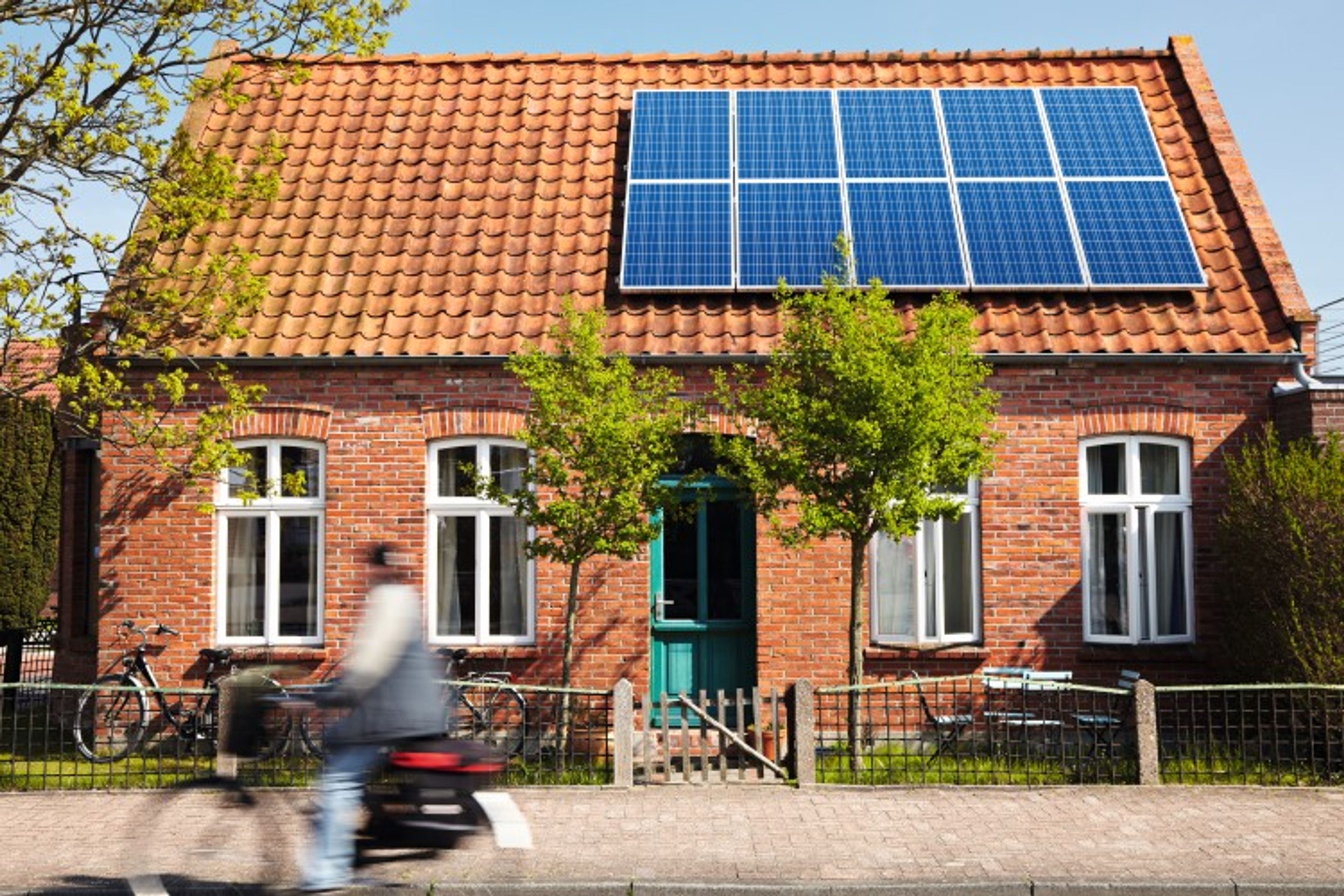 Huis met 1 verdieping: 2 ramen , een deur, 2 ramen. En zonnepanelen op het dak. Fietser fietst voorbij