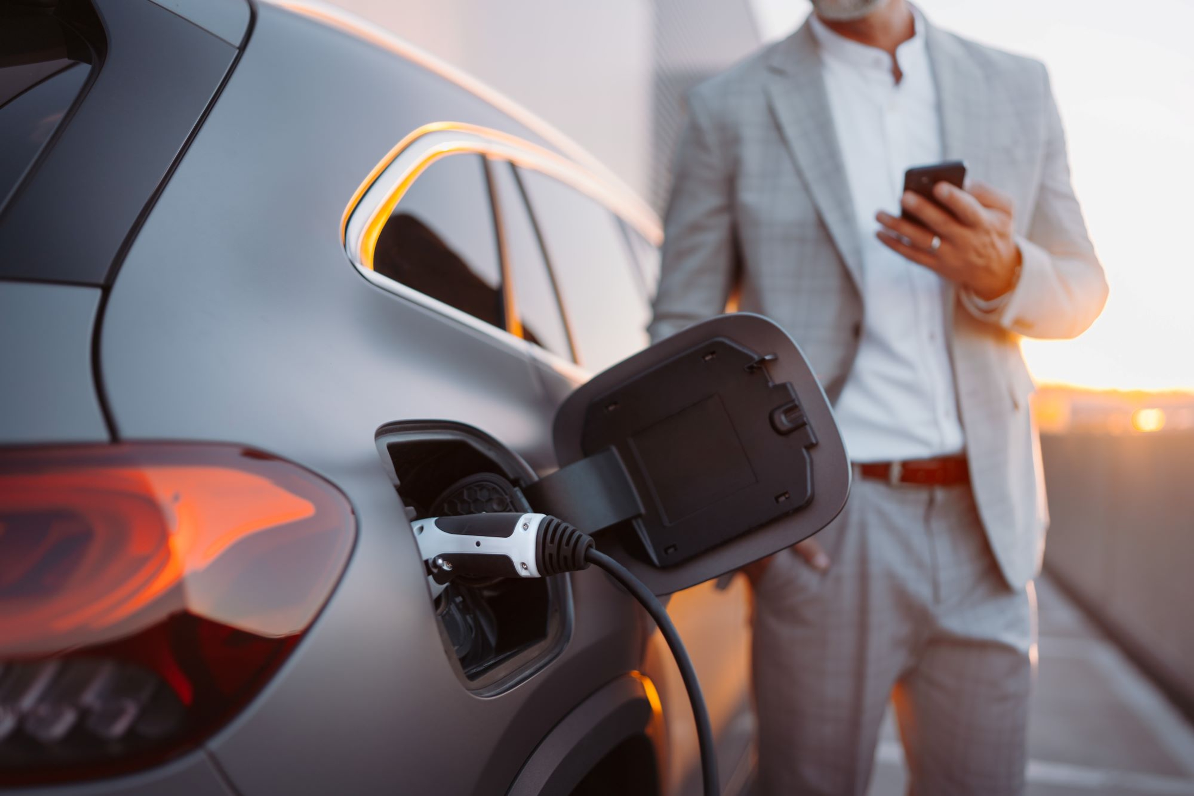 Man waarvan we het hoofd niet zien, kijkt op smartphone bij zijn wagen die staat te laden