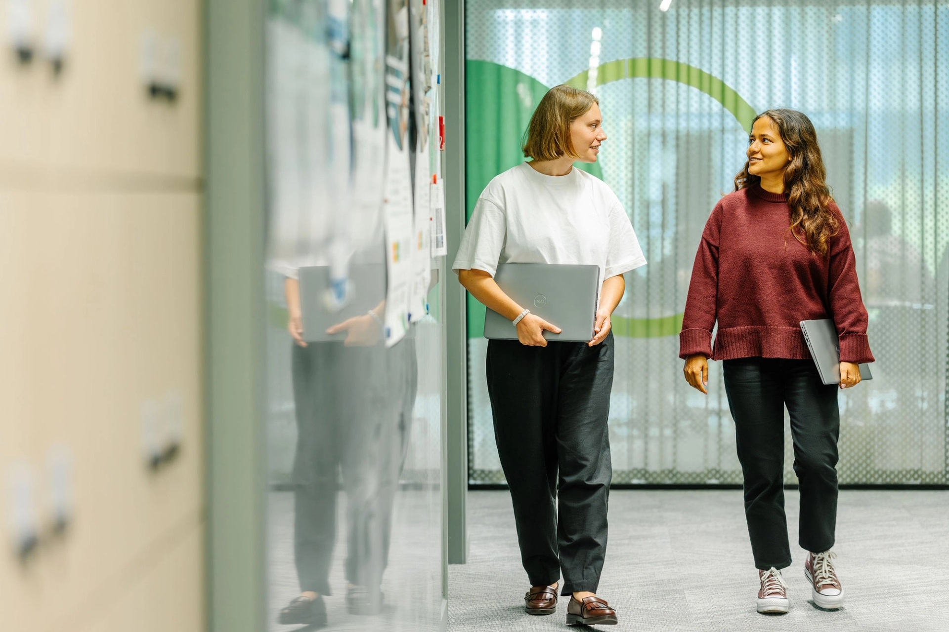 Deux femmes marchent dans le couloir d'un bureau, chacune tenant un ordinateur portable. Elles sourient et discutent, créant une atmosphère amicale et professionnelle.