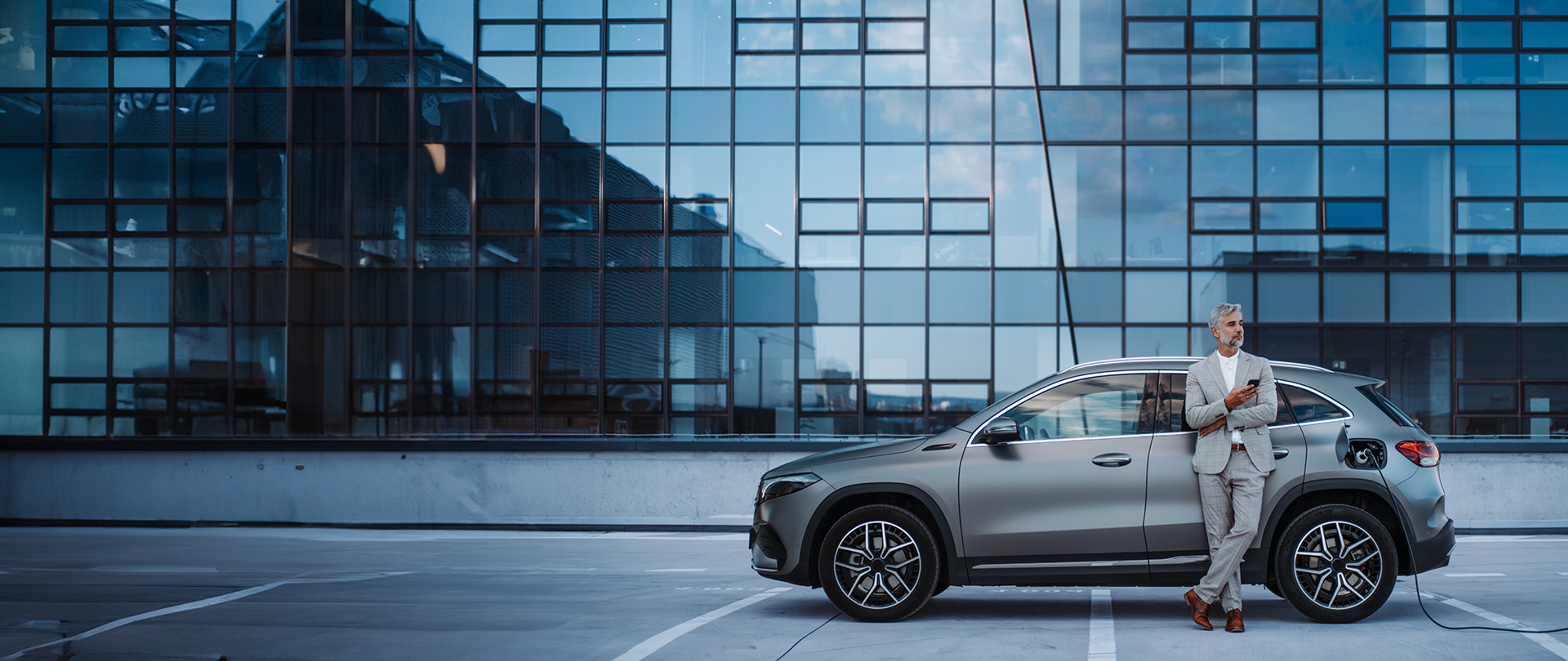 Elektrische wagen is aan het opladen voor modern gebouw met veel glas. Man in pak staat erbij en kijkt op gsm.