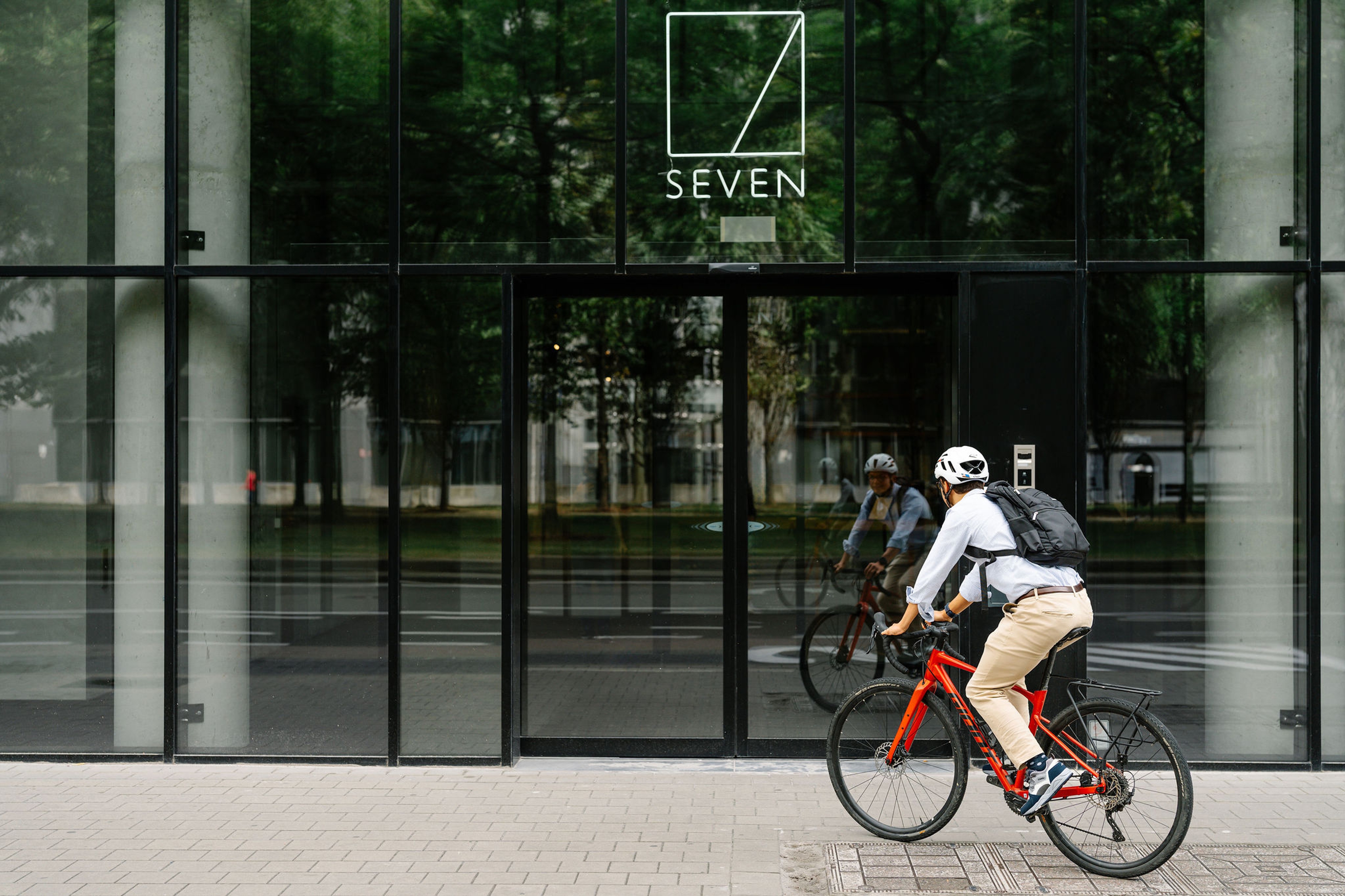 Un cycliste portant un casque et un sac à dos roule sur un vélo rouge devant un bâtiment moderne aux murs de verre élégants. Le reflet montre les arbres et les rues de la ville.