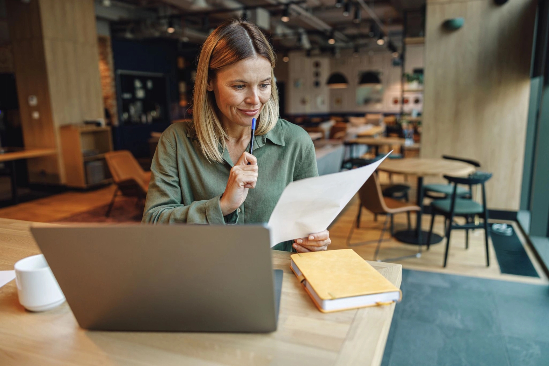 Une femme portant un t-shirt vert est assise à une table de café conviviale. Elle parcourt un document, un stylo à la main. Un ordinateur portable et un carnet se trouvent à proximité.