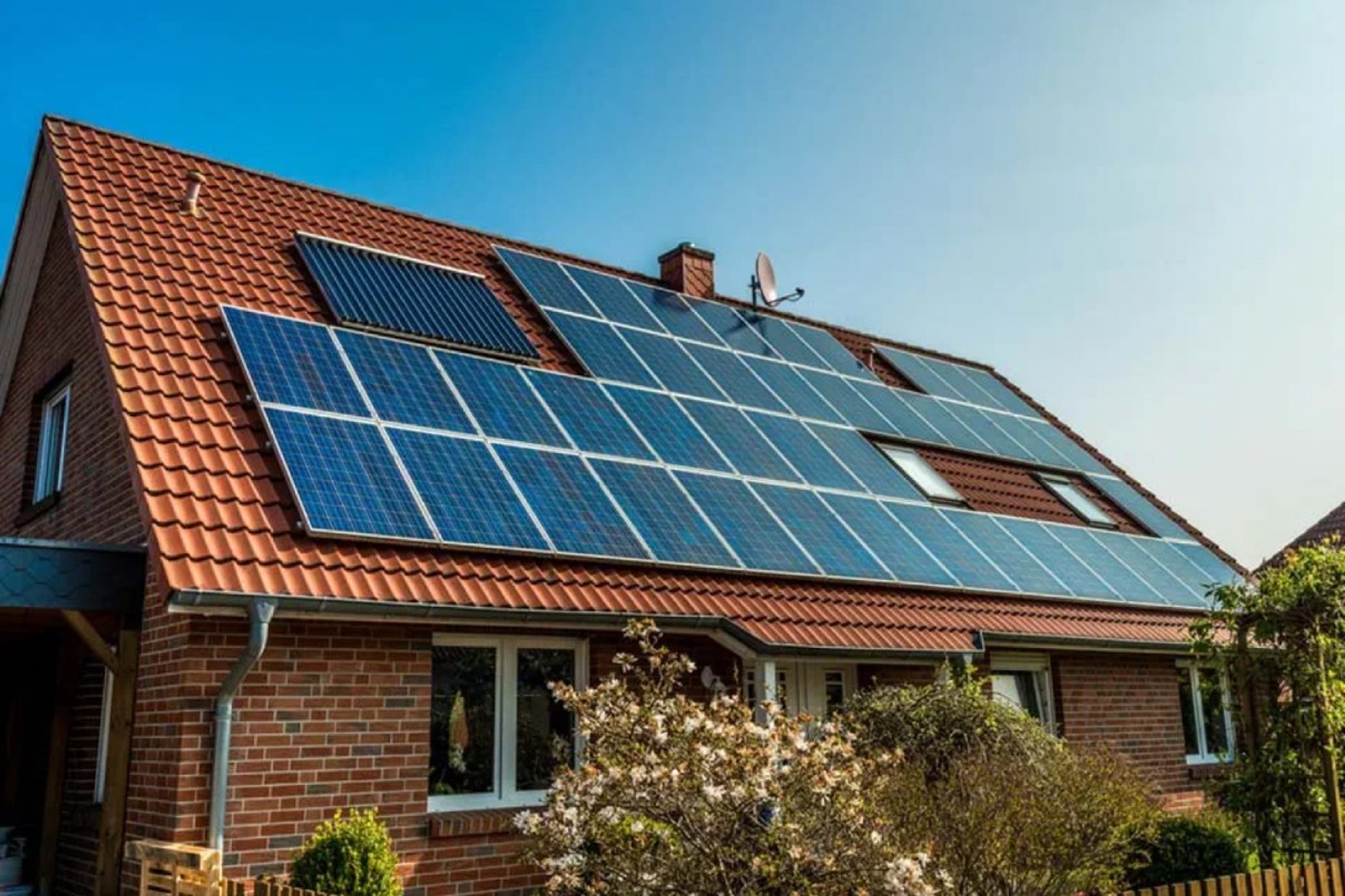 Huis met zonnepanelen op het dak, struiken op de voorgrond, blauwe lucht.