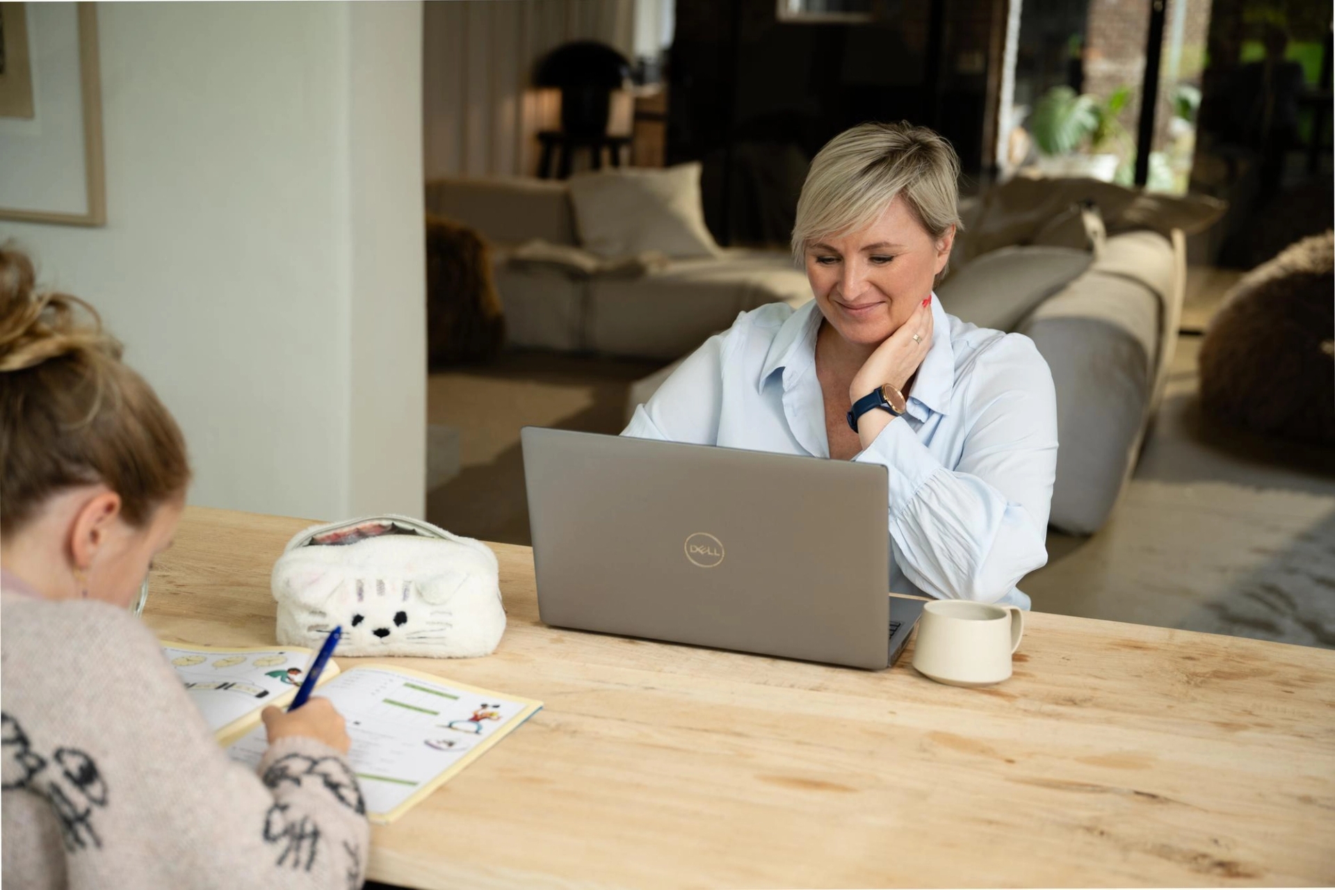 Een vrouw met kort blond haar glimlacht terwijl ze op een laptop werkt aan een houten tafel. Een kind zit tegenover haar te tekenen in een gezellige, goed verlichte kamer.