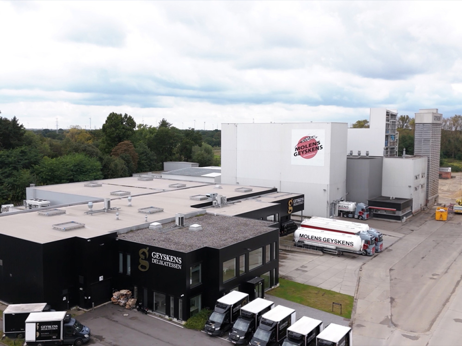 Vue aérienne du site industriel de Molens Geyskens avec plusieurs bâtiments modernes, des camions de transport stationnés et des véhicules utilitaires devant les locaux, entouré de verdure