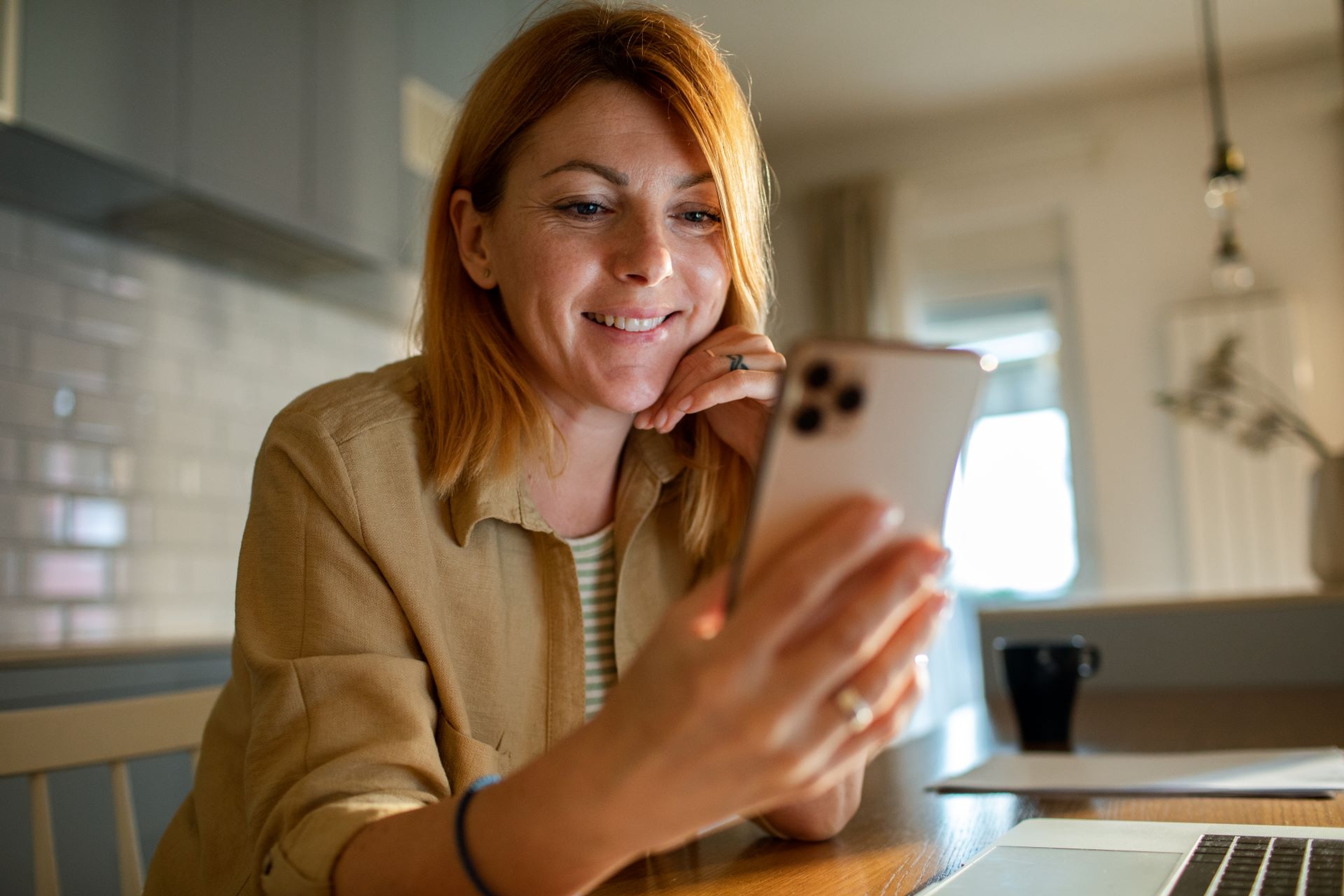Une femme souriante assise à une table regarde son smartphone, avec un ordinateur portable devant elle dans une cuisine
