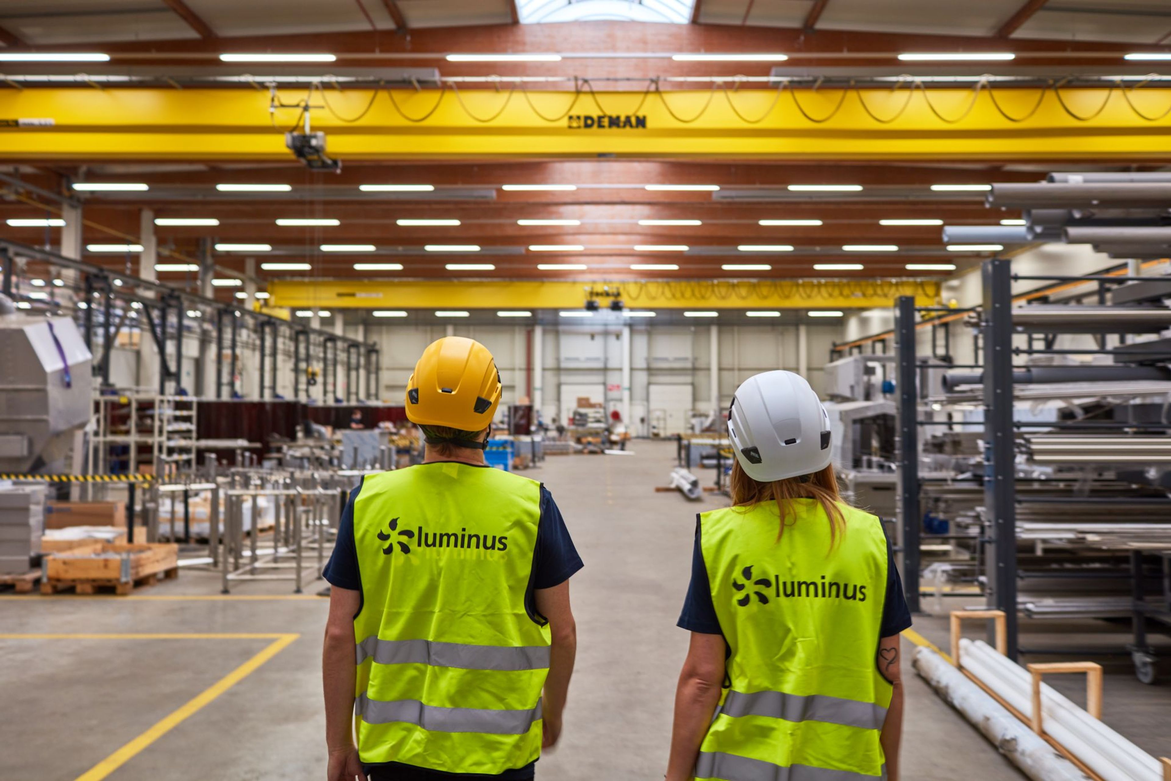 Twee werknemers met veiligheidsvesten en helmen staan in een helder verlichte fabrieksgang. Boven hun hoofden is een gele kraan te zien. De scène is ordelijk en geconcentreerd.