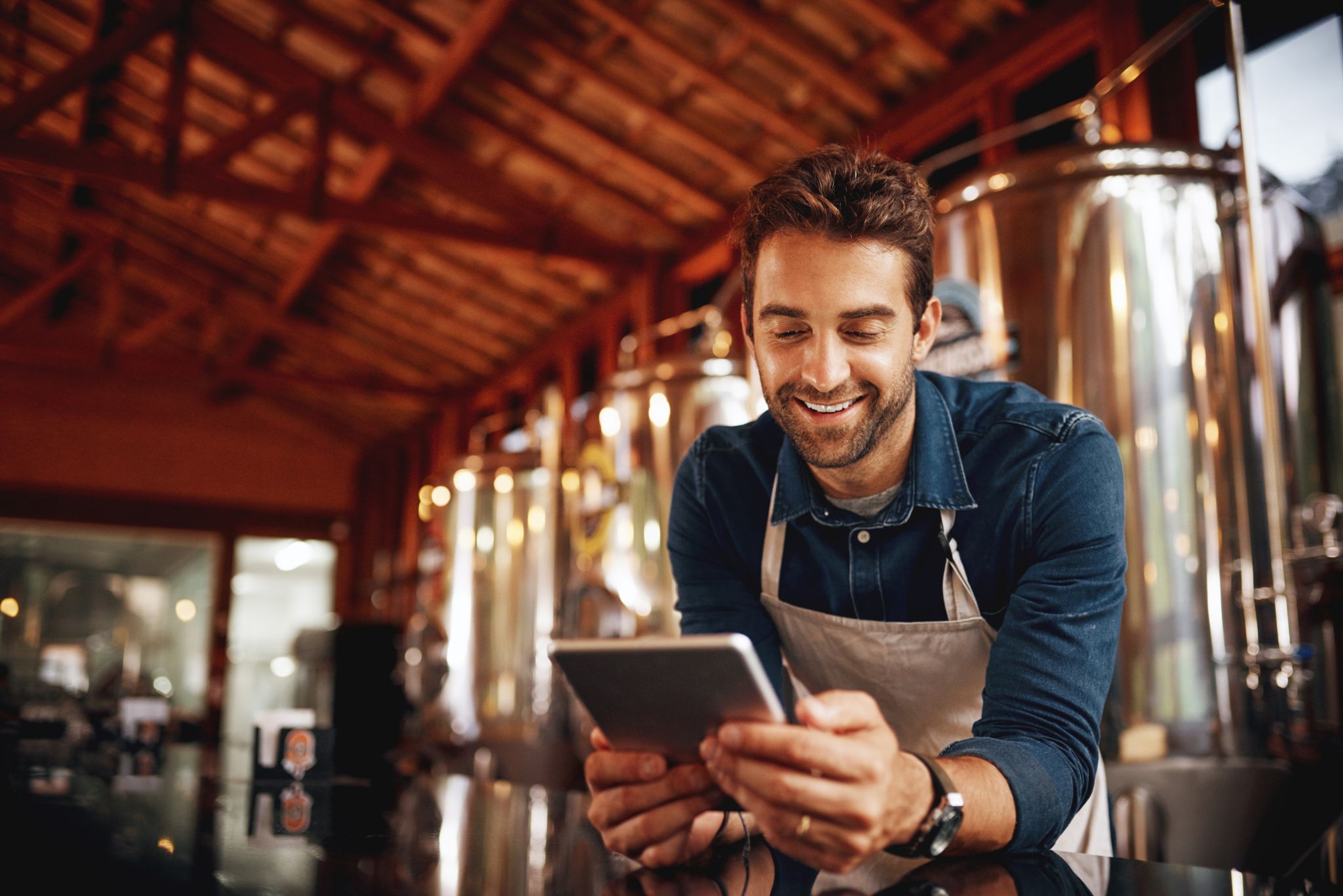 Un homme portant une chemise en jean et un tablier regarde une tablette avec le sourire dans une brasserie, entouré de grandes cuves en acier inoxydable et d’une charpente en bois apparente