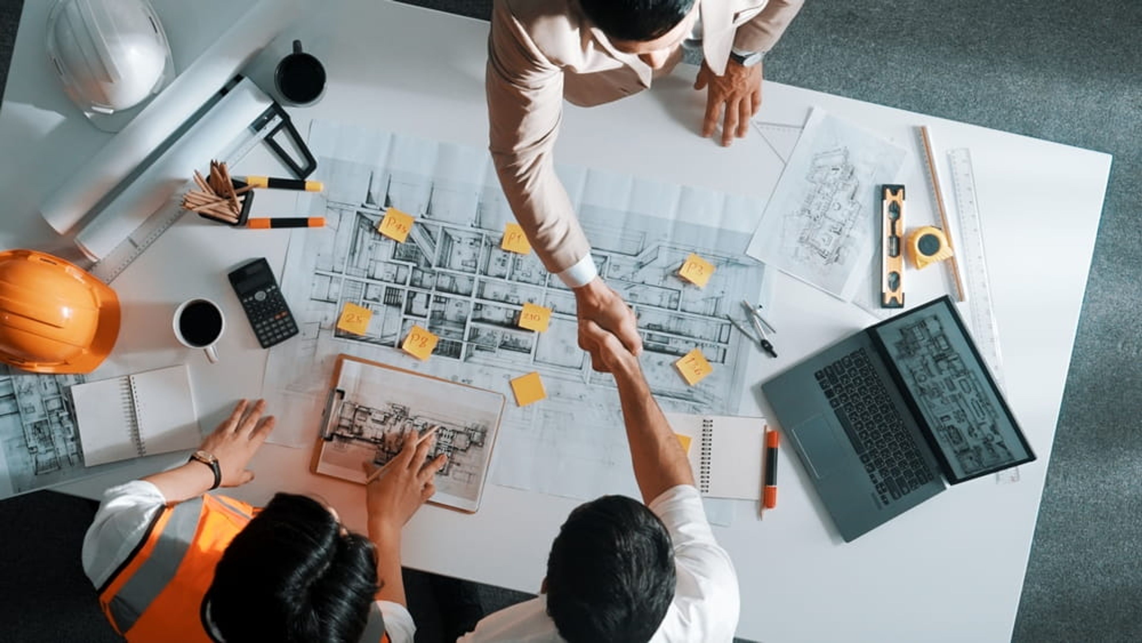 Bovenaanzicht van architectenplan, 3 mensen waarvan 2 de hand schudden.