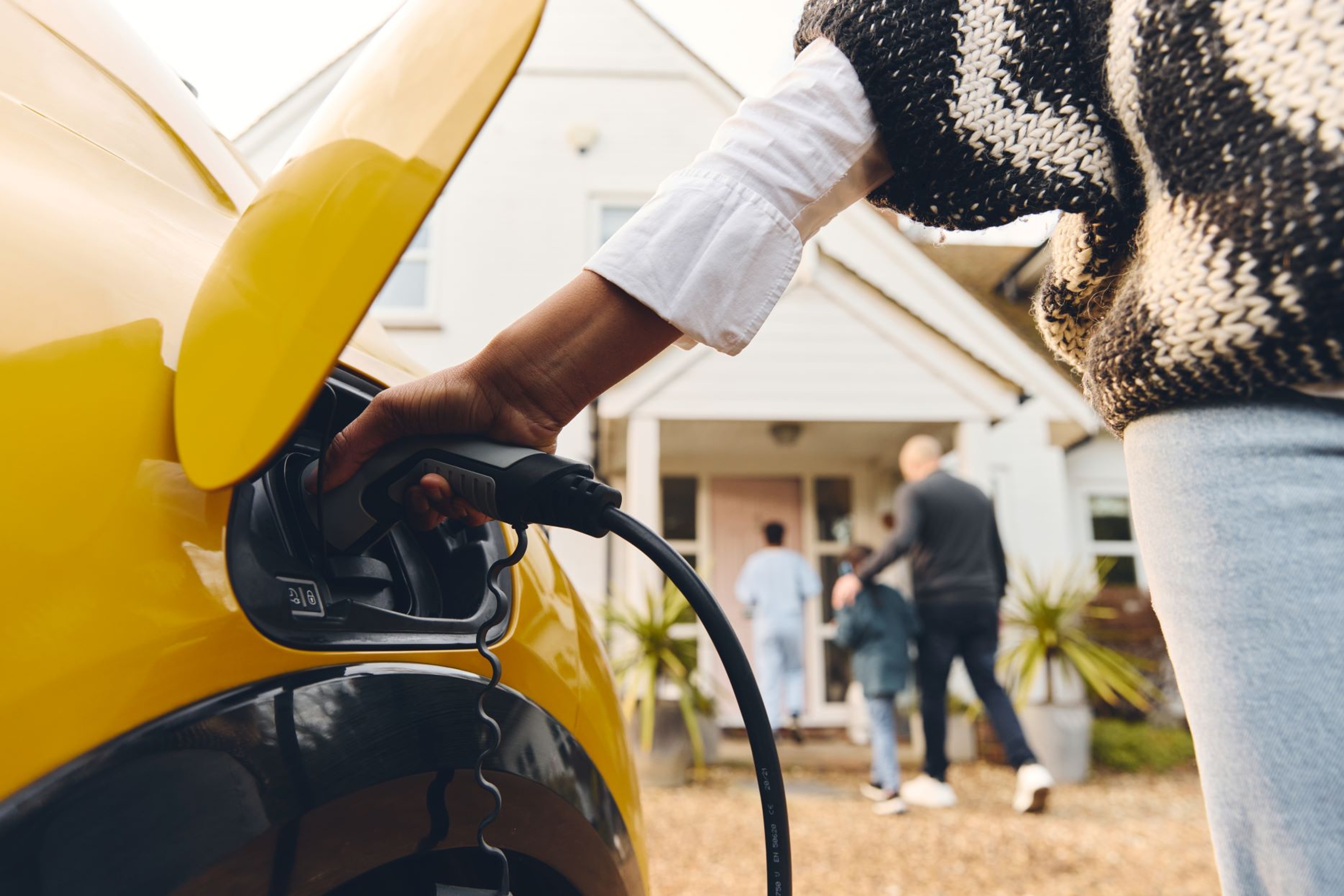 Une femme branche sa voiture électrique pour une recharge à domicile, pendant qu'un homme et deux enfants rentrent dans la maison