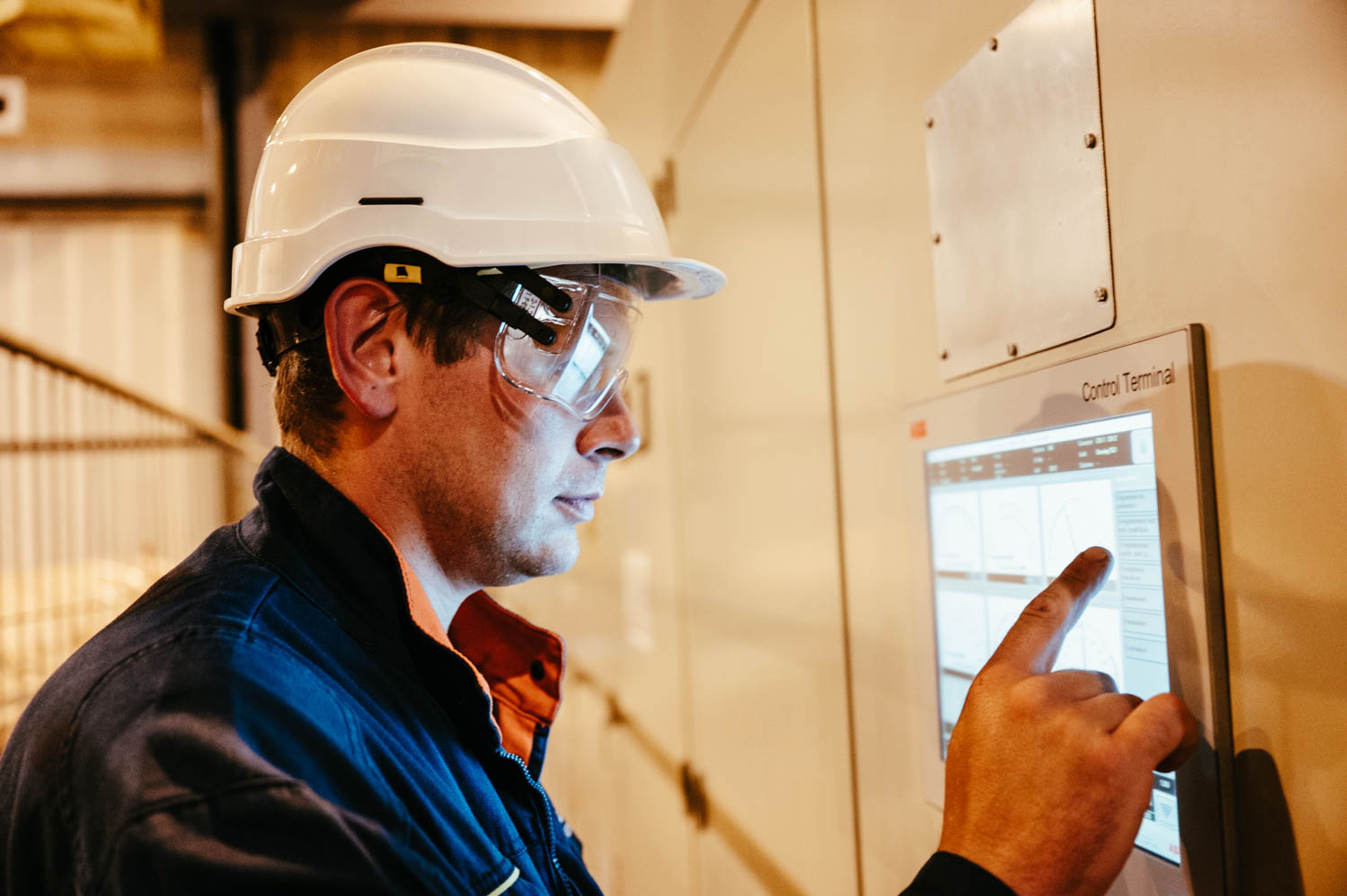 Un ouvrier portant un casque blanc et des lunettes de sécurité interagit avec un écran de contrôle. Le décor est industriel, suggérant la concentration et l'attention.