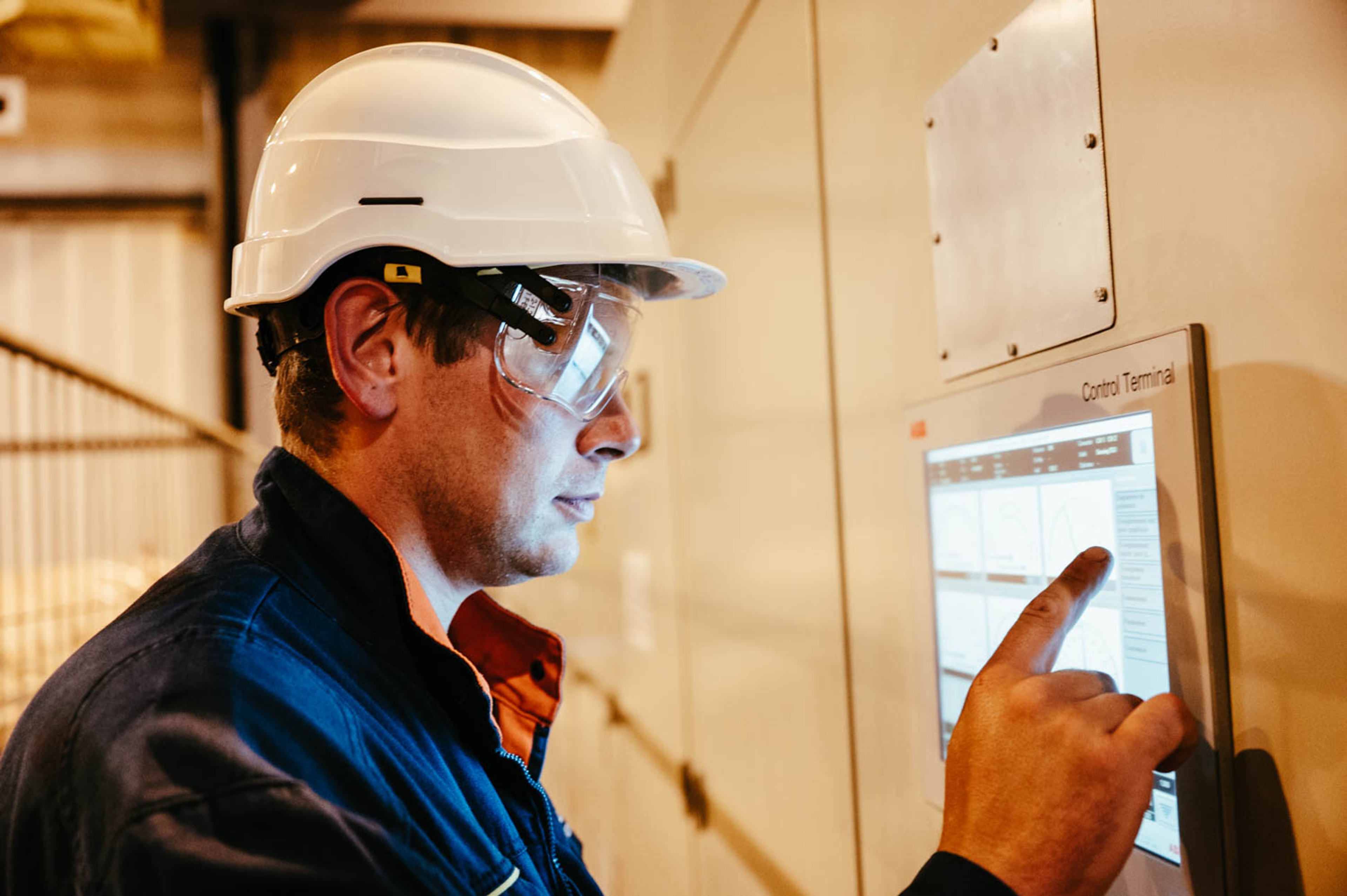 Un ouvrier portant un casque blanc et des lunettes de sécurité interagit avec un écran de contrôle. Le décor est industriel, suggérant la concentration et l'attention.