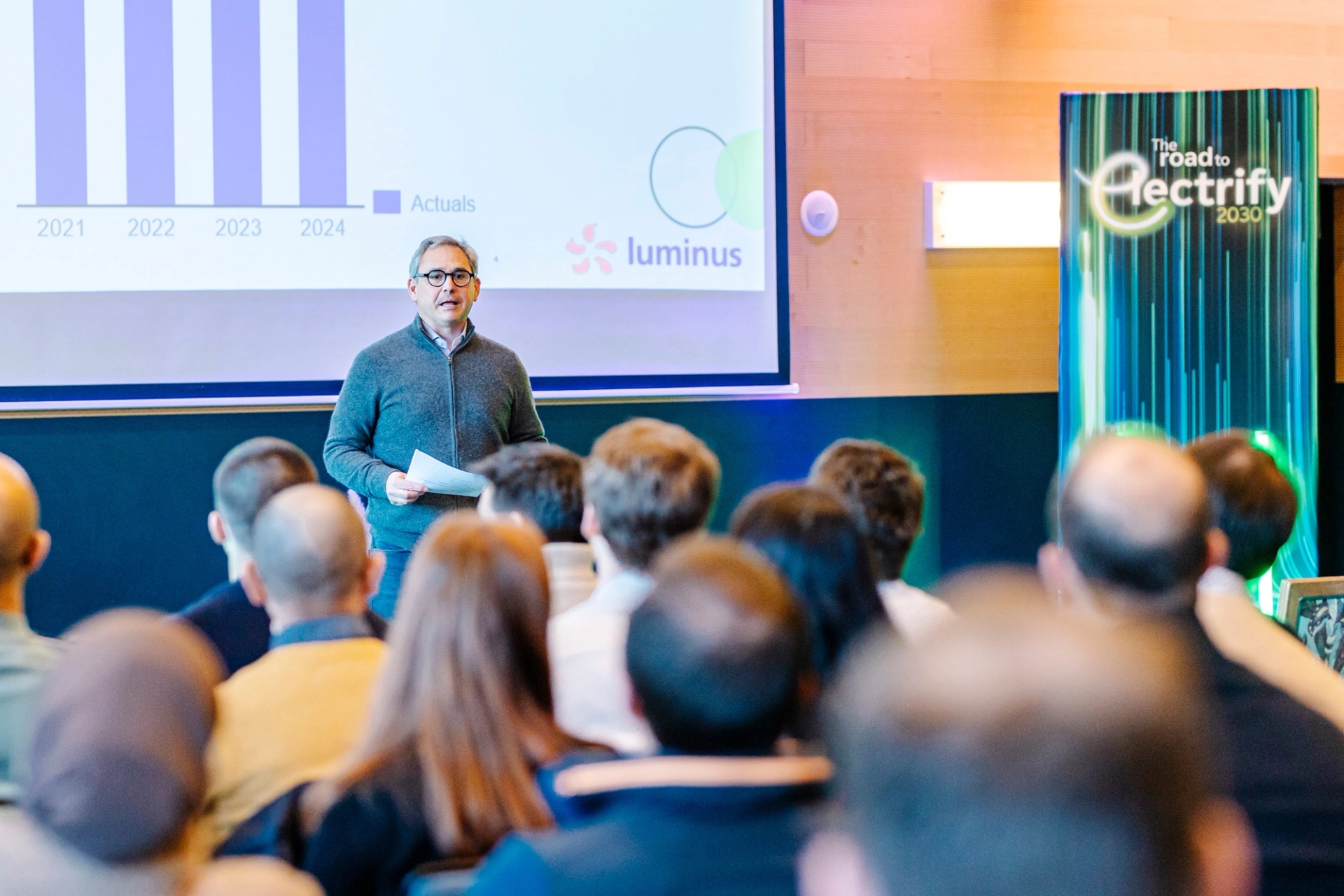Een spreker staat voor een publiek en presenteert een grafiek op een scherm met de titel ‘Luminus’ met gegevens van 2021 tot 2024. De setting is een vergaderzaal met een banner met de tekst ‘The Road to Electrify 2030’.