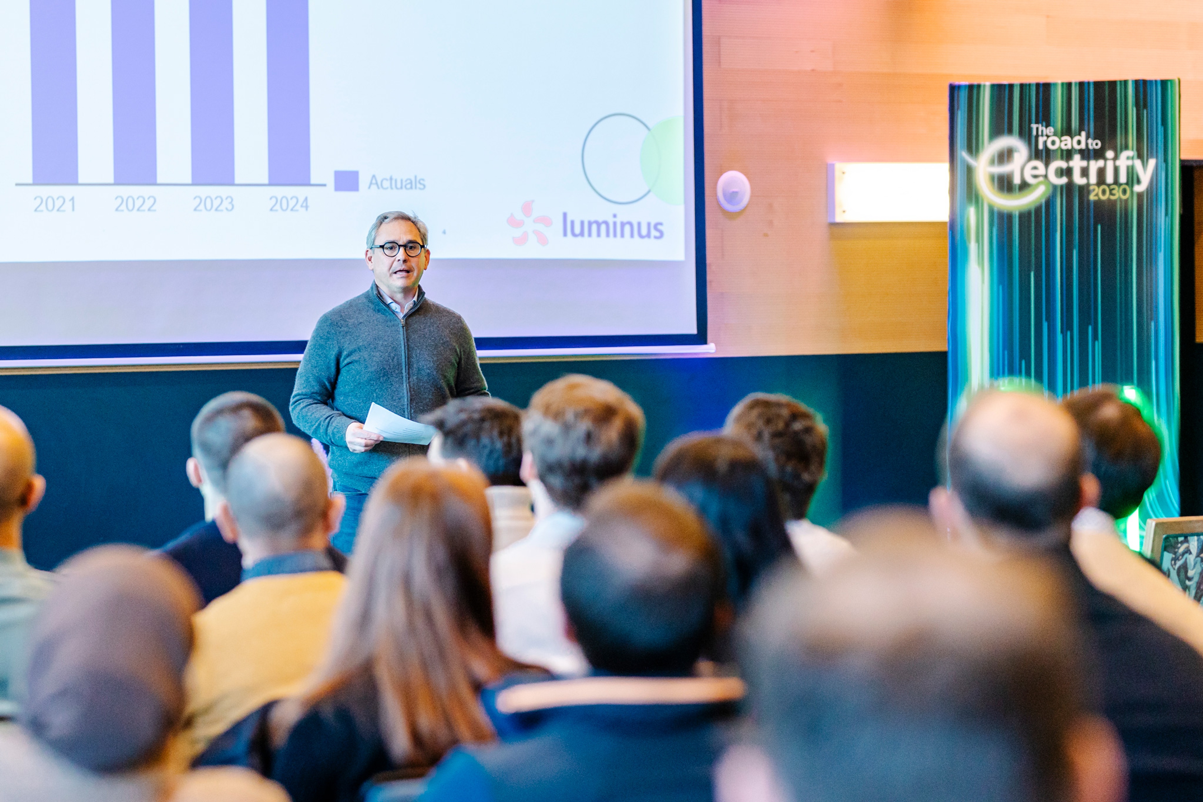 Een spreker staat voor een publiek en presenteert een grafiek op een scherm met de titel ‘Luminus’ met gegevens van 2021 tot 2024. De setting is een vergaderzaal met een banner met de tekst ‘The Road to Electrify 2030’.