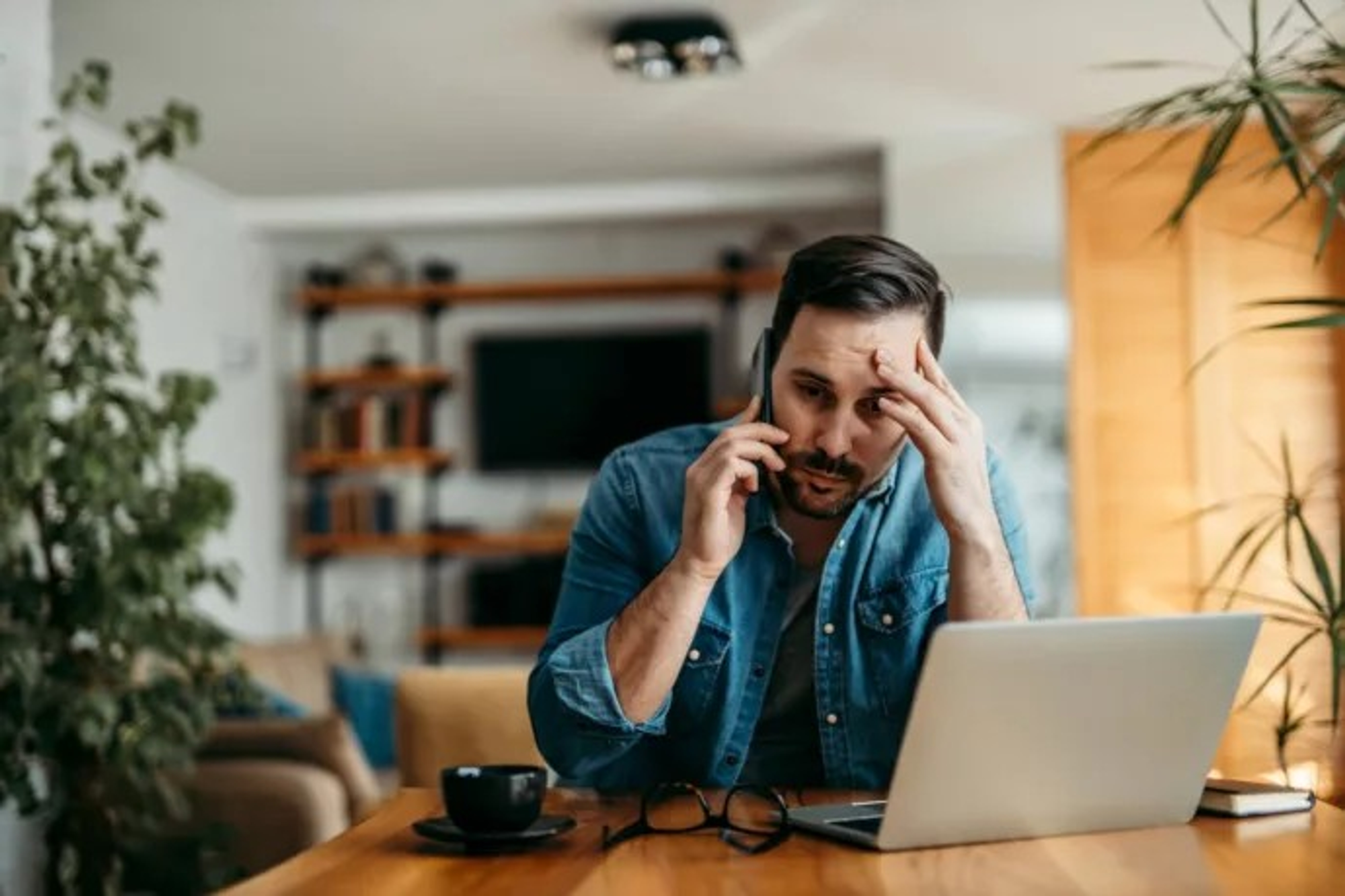 Man met jeanshemd in woonkamer zit te bellen, hand aan hoofd, laptop op tafel.