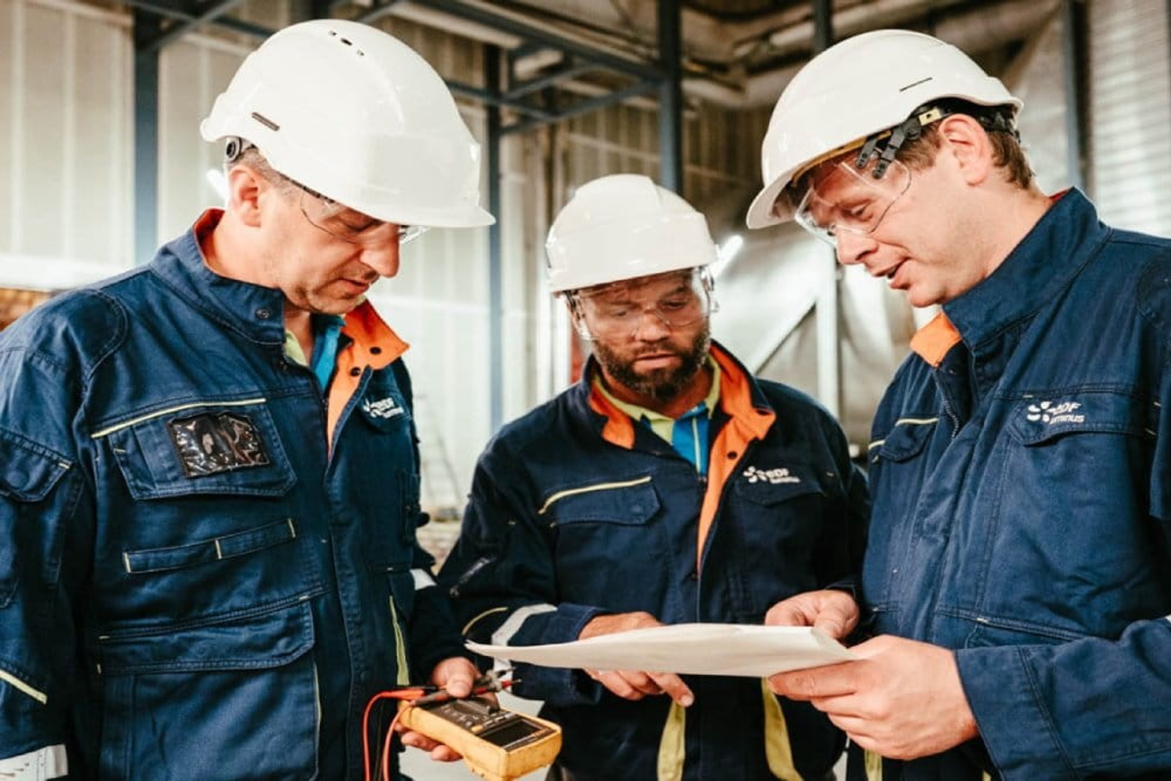 Trois techniciens en tenue de travail et casque de sécurité discutent dans un environnement industriel