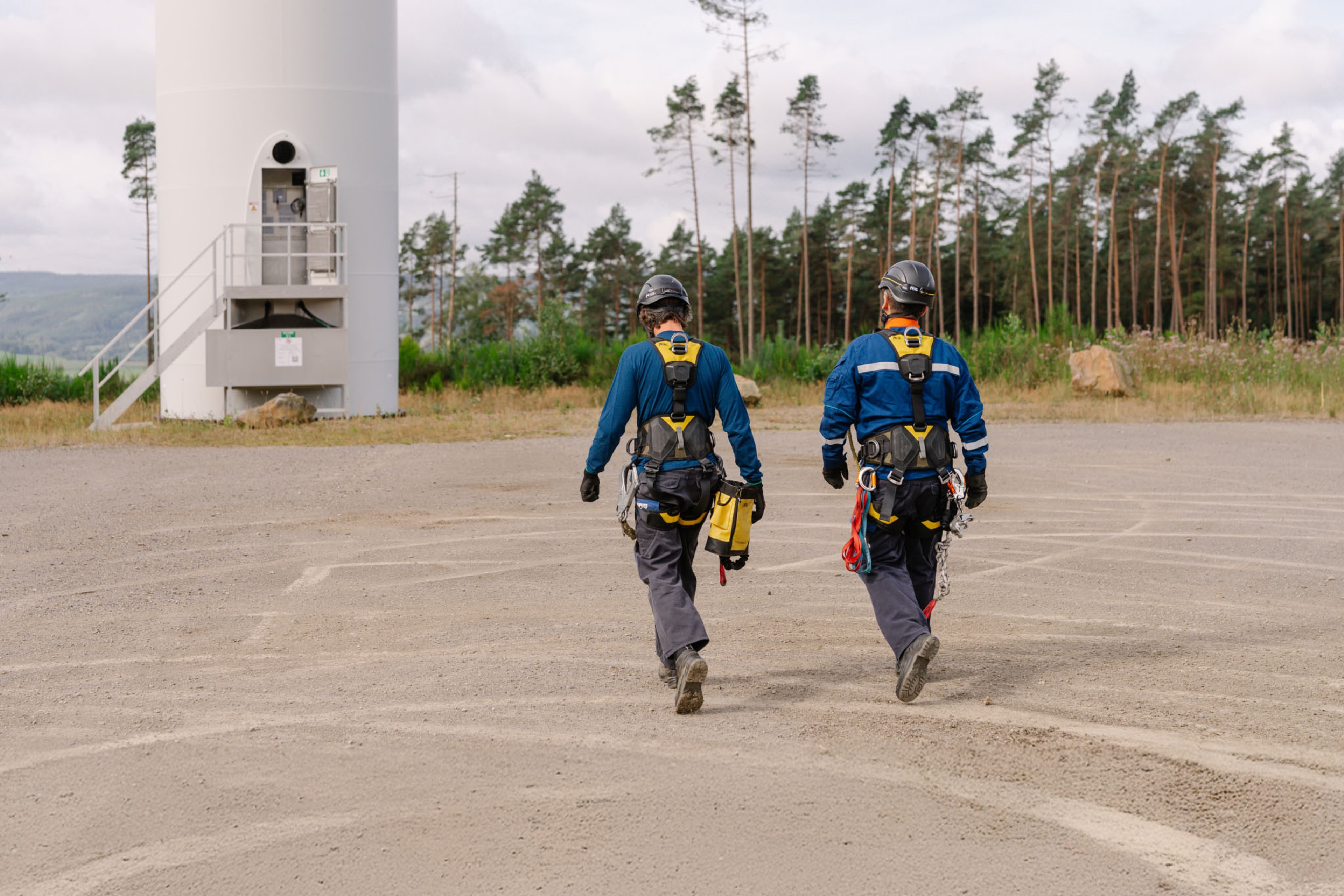 Deux techniciens en tenue de sécurité marchent vers la base d’une éolienne, équipés de harnais et d’outils.