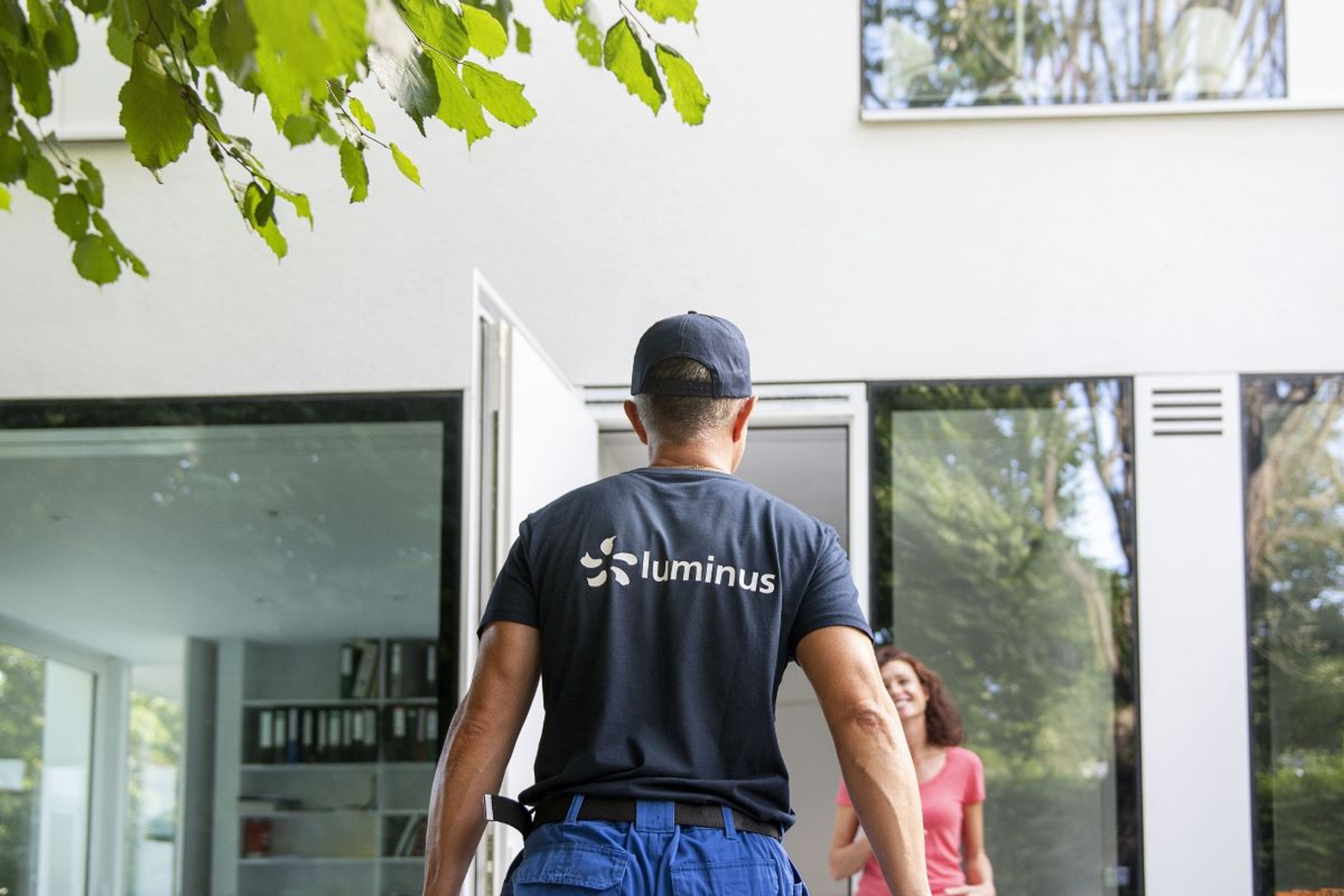 Technieker van Luminus komt aan bij wit modern huis. Dame met rode top wacht hem lachend op bij de open voordeur.