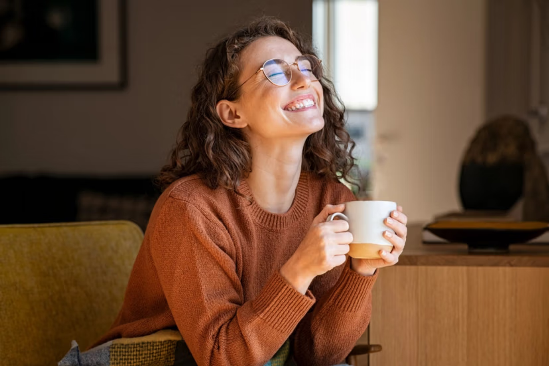 Jongedame zit in de living met kopje in de hand, gelukzalige smile en gesloten ogen.