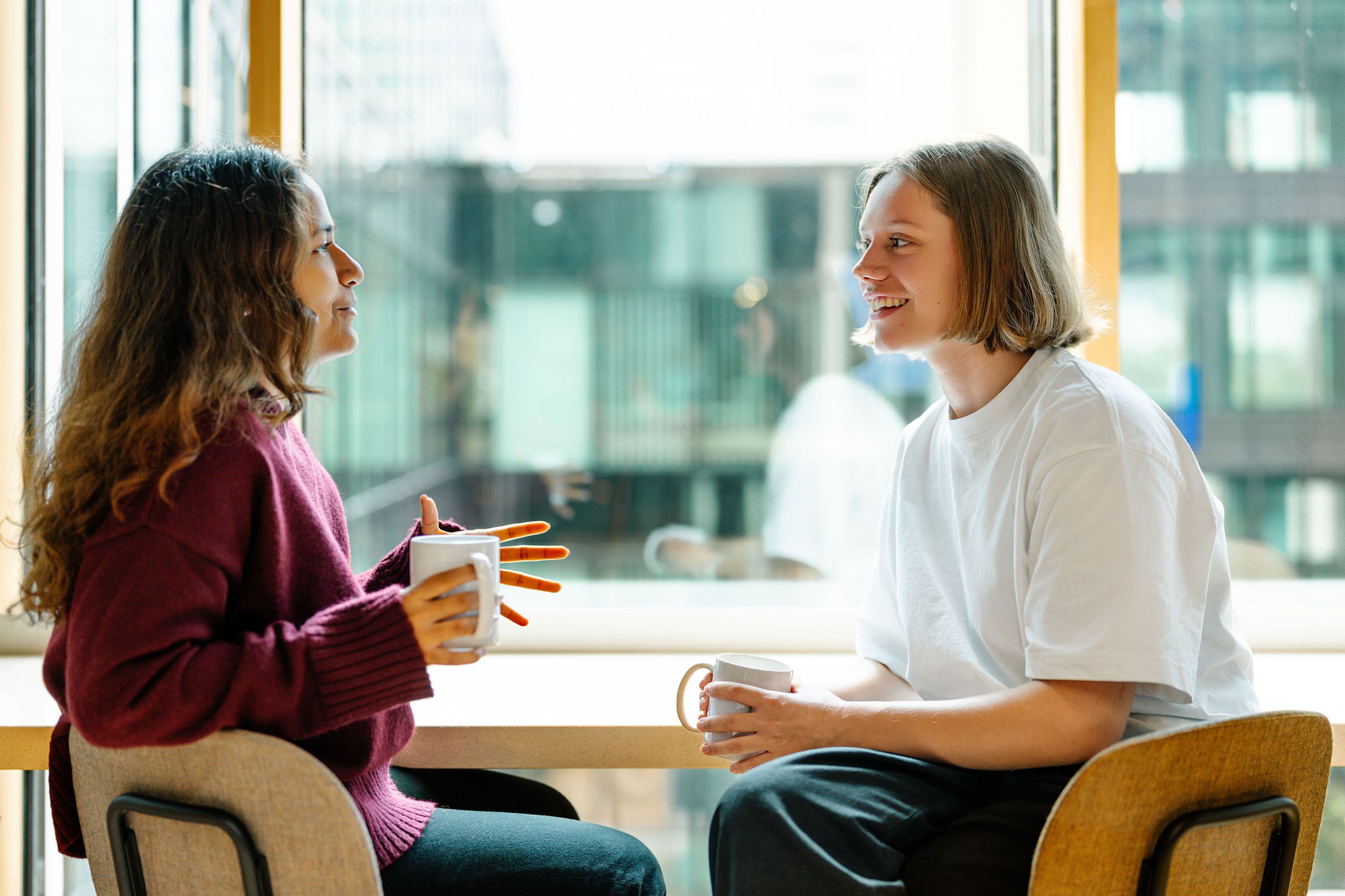 Two people sit at a table by a window, smiling and holding mugs. They seem engaged in a friendly conversation, creating a warm, relaxed atmosphere.