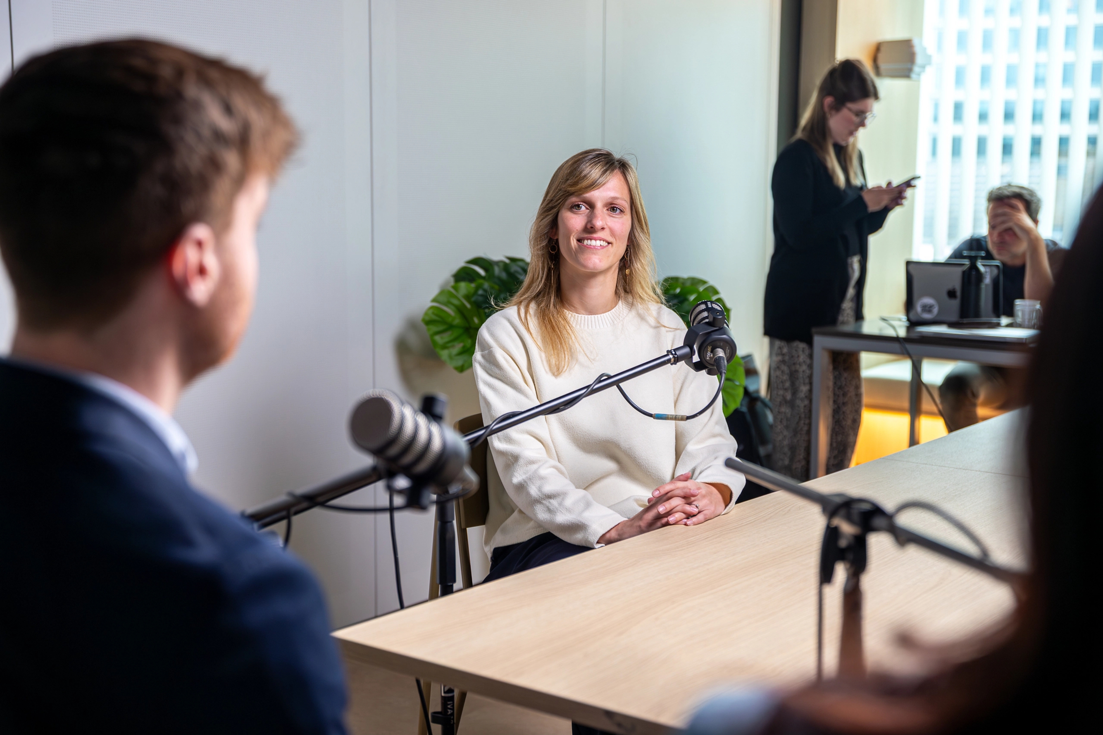 Opname van webinar: dame met witte trui achter de microfoon, man zien we van op de rug. In de achtergrond 2 mensen bezig met de techniek.