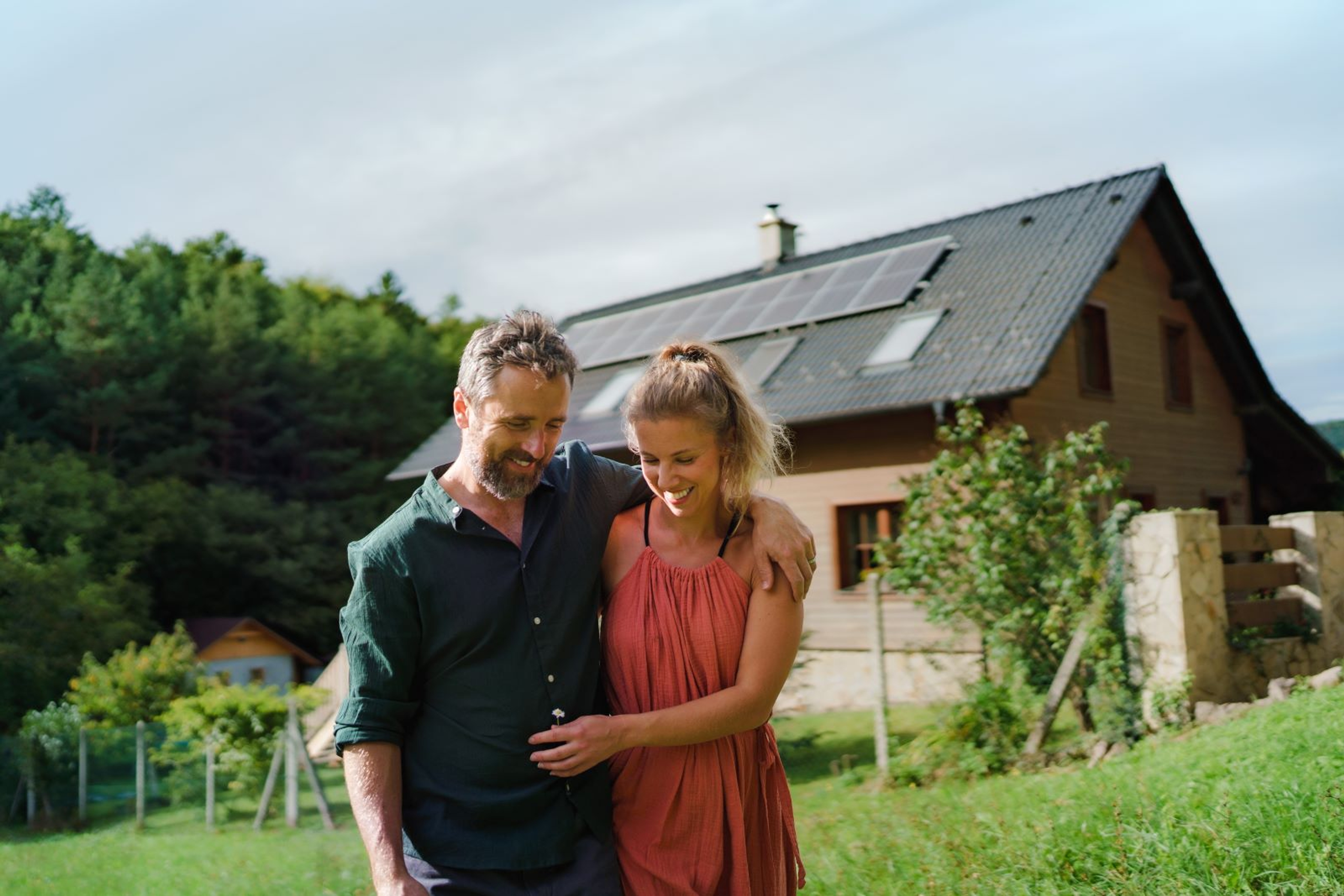 Couple souriant marchant bras dessus bras dessous dans un jardin verdoyant, devant une maison équipée de panneaux solaires sur le toit