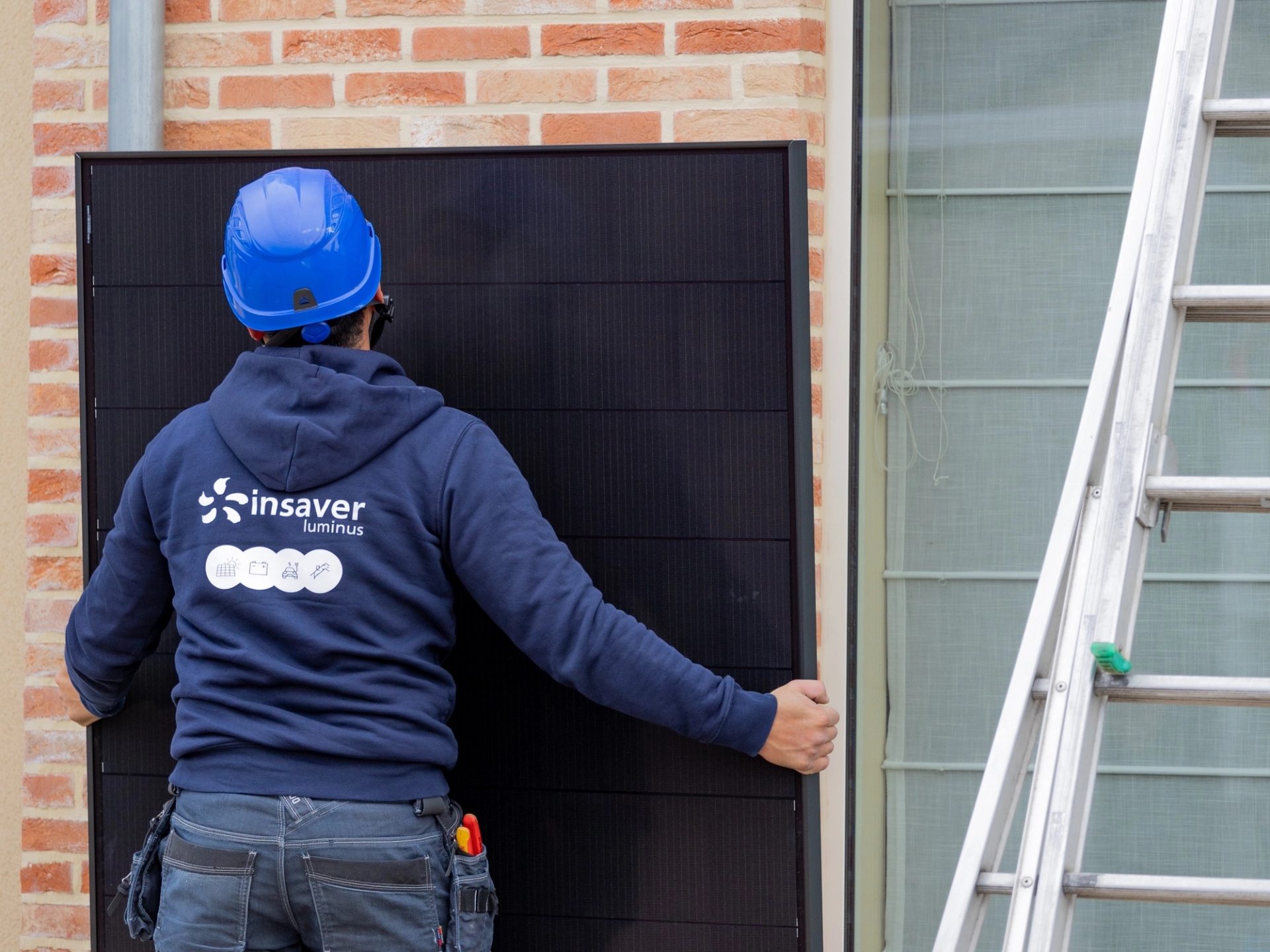 Technieker met Insaver hoodie en blauwe helm zet zonnepaneel tegen de muur van een villa, ladder rechts.