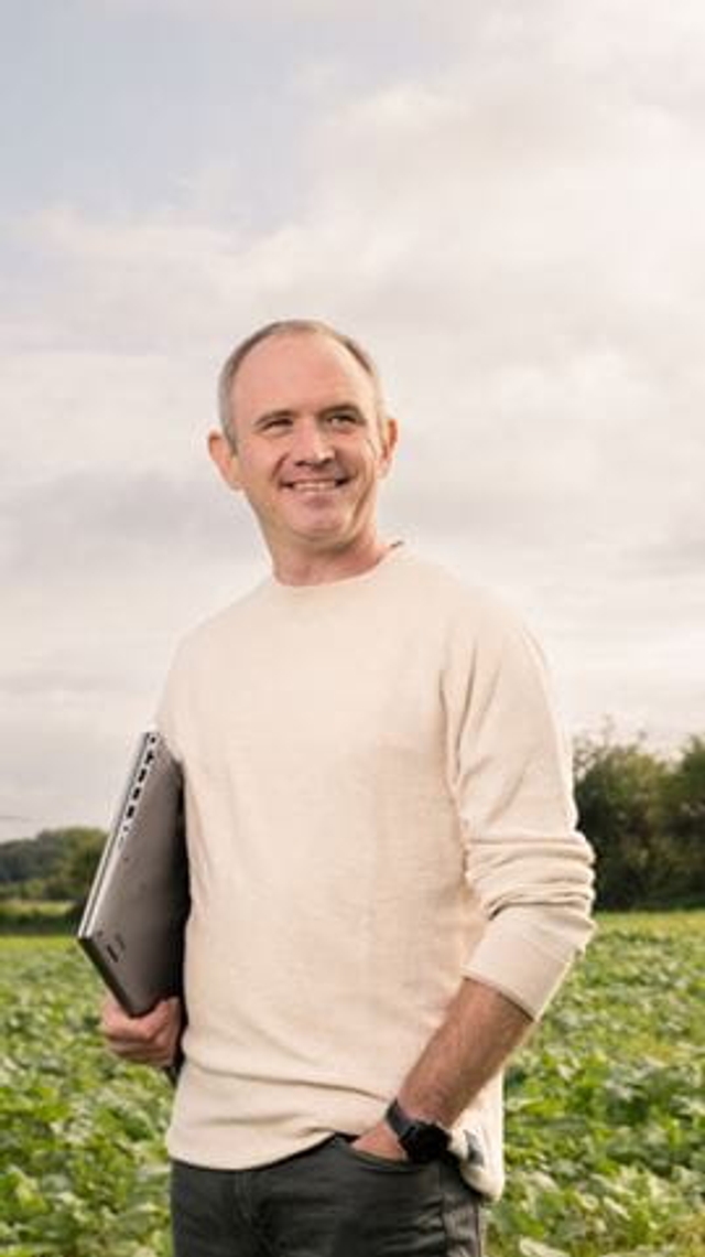 Kim holding his laptop, stands before a green field.