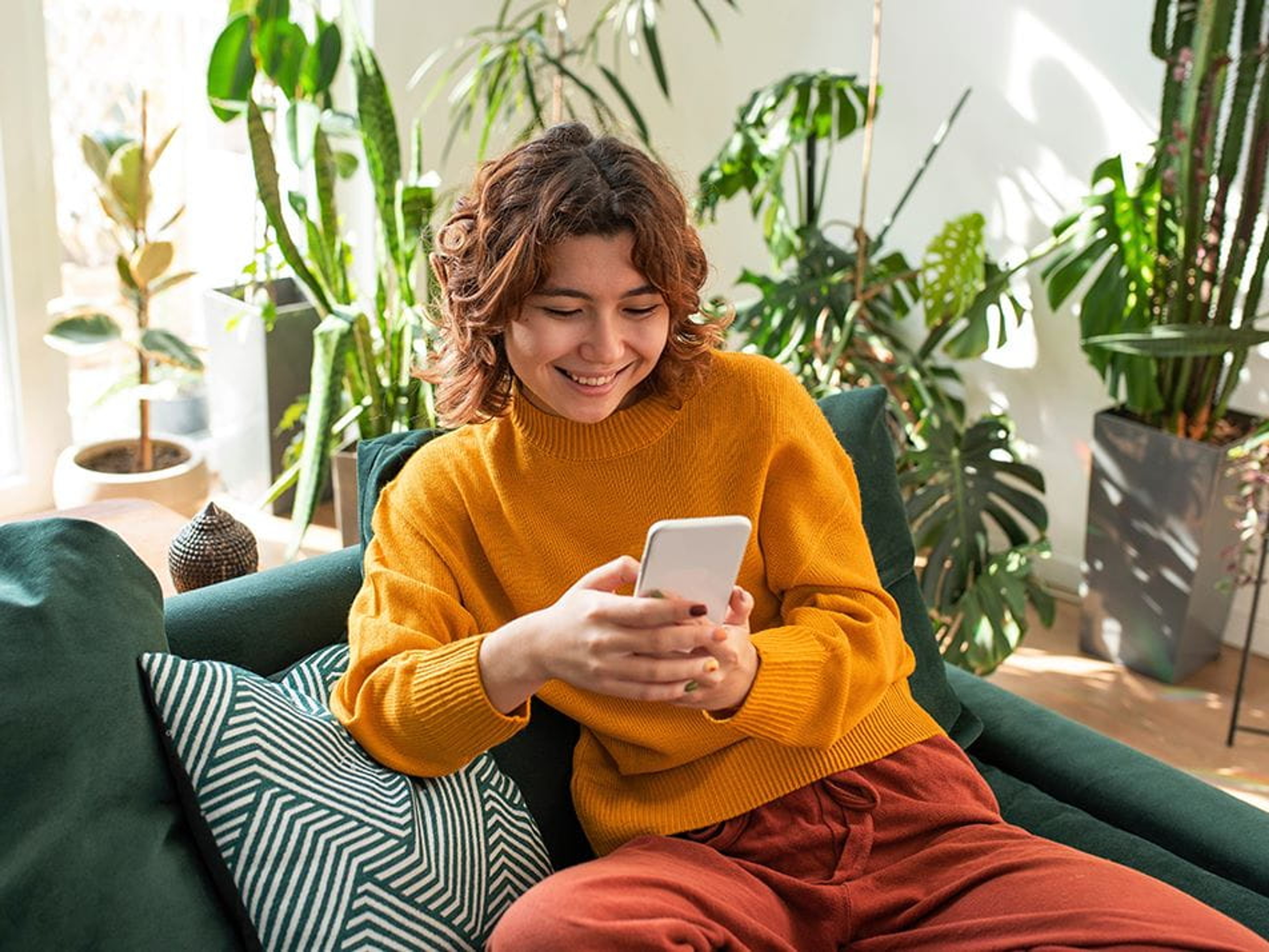 Jonge vrouw lachend in de zetel, kijkt op smartphone. oranje trui, rode broek, gatenplant