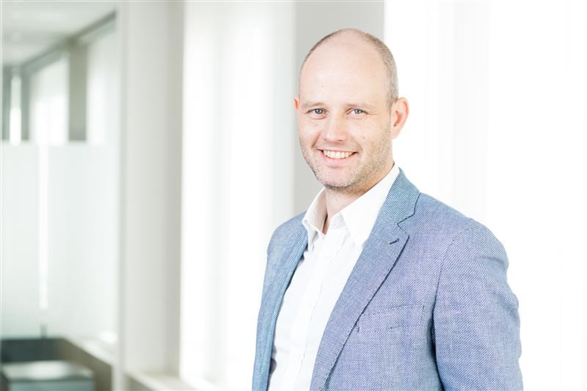 Portrait d’un homme souriant en veste bleue claire, debout dans un couloir lumineux