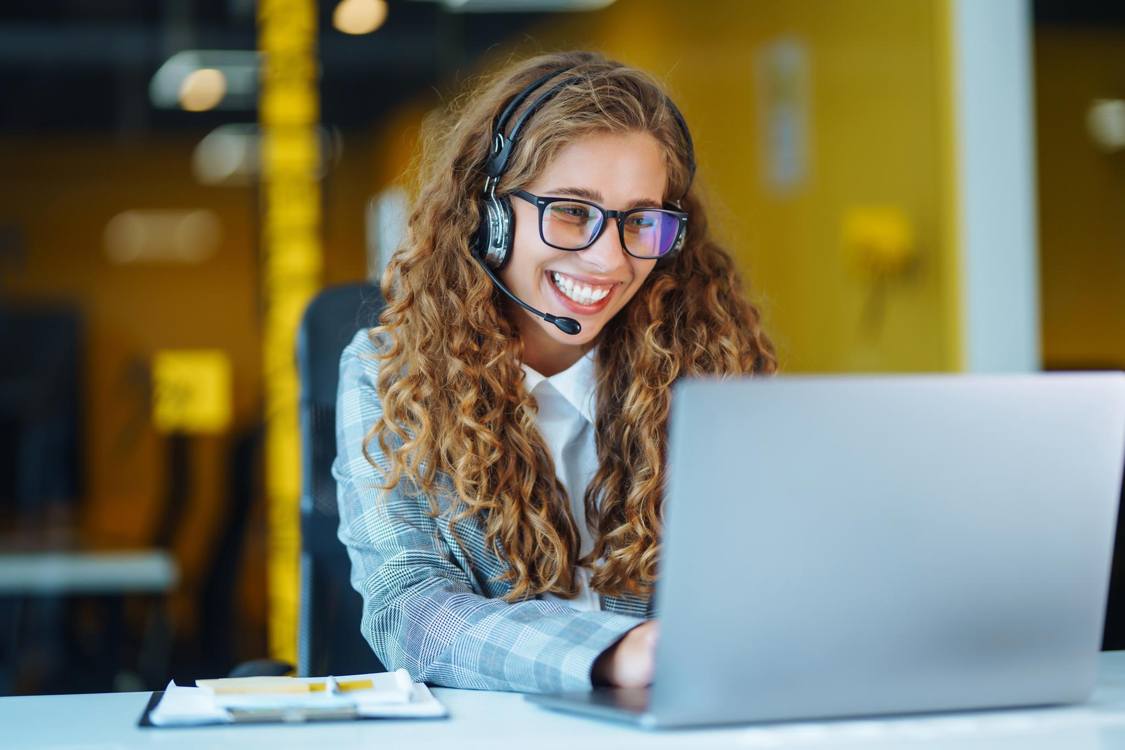 Une femme aux cheveux bouclés, portant des lunettes et un casque audio, sourit tout en travaillant sur un ordinateur portable dans un bureau lumineux et moderne, créant une ambiance engageante et joyeuse.