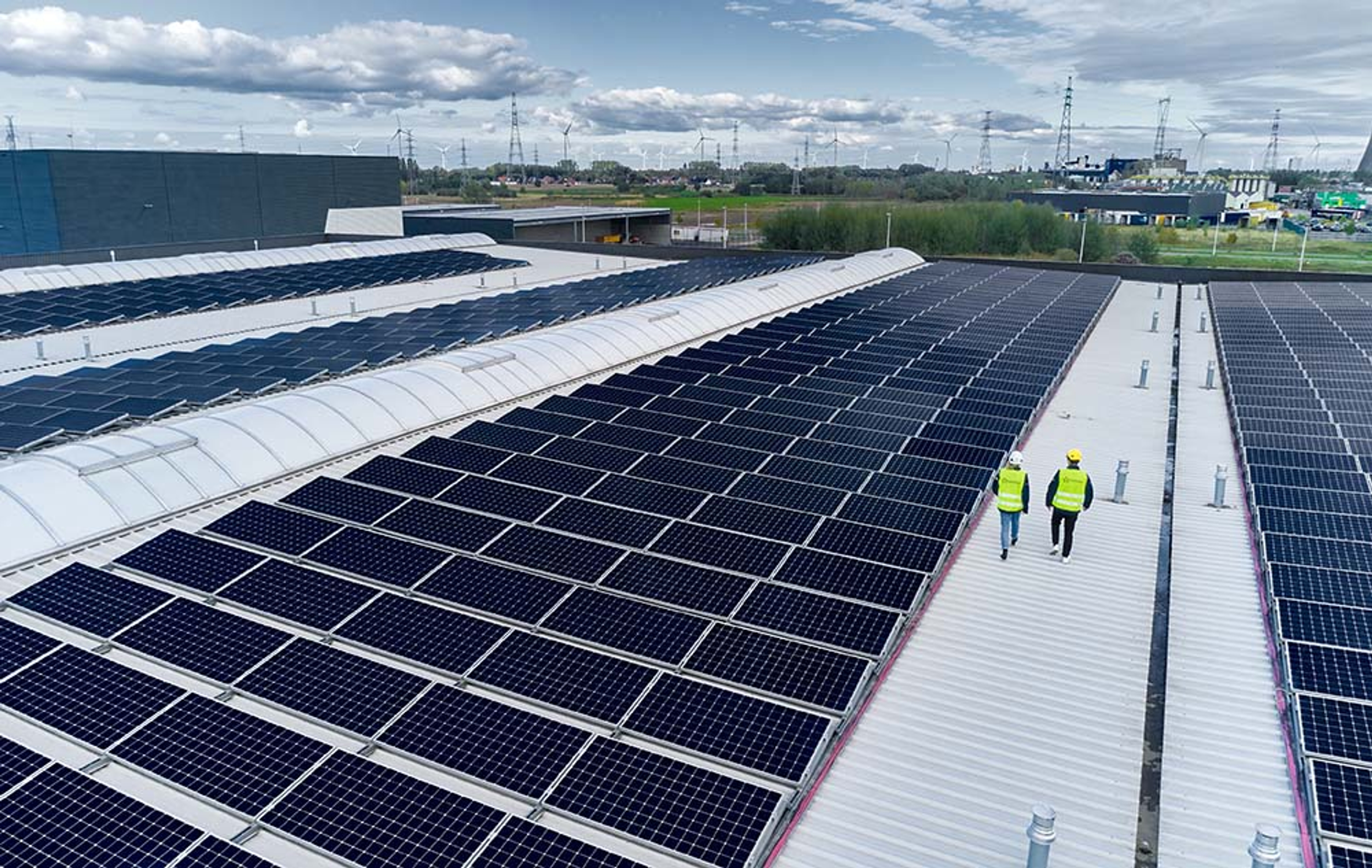 Des panneaux solaires installés sur le toit d'un grand bâtiment industriel. Deux techniciens en gilet jaune et casque de sécurité marchent entre les rangées de panneaux, sous un ciel partiellement nuageux.