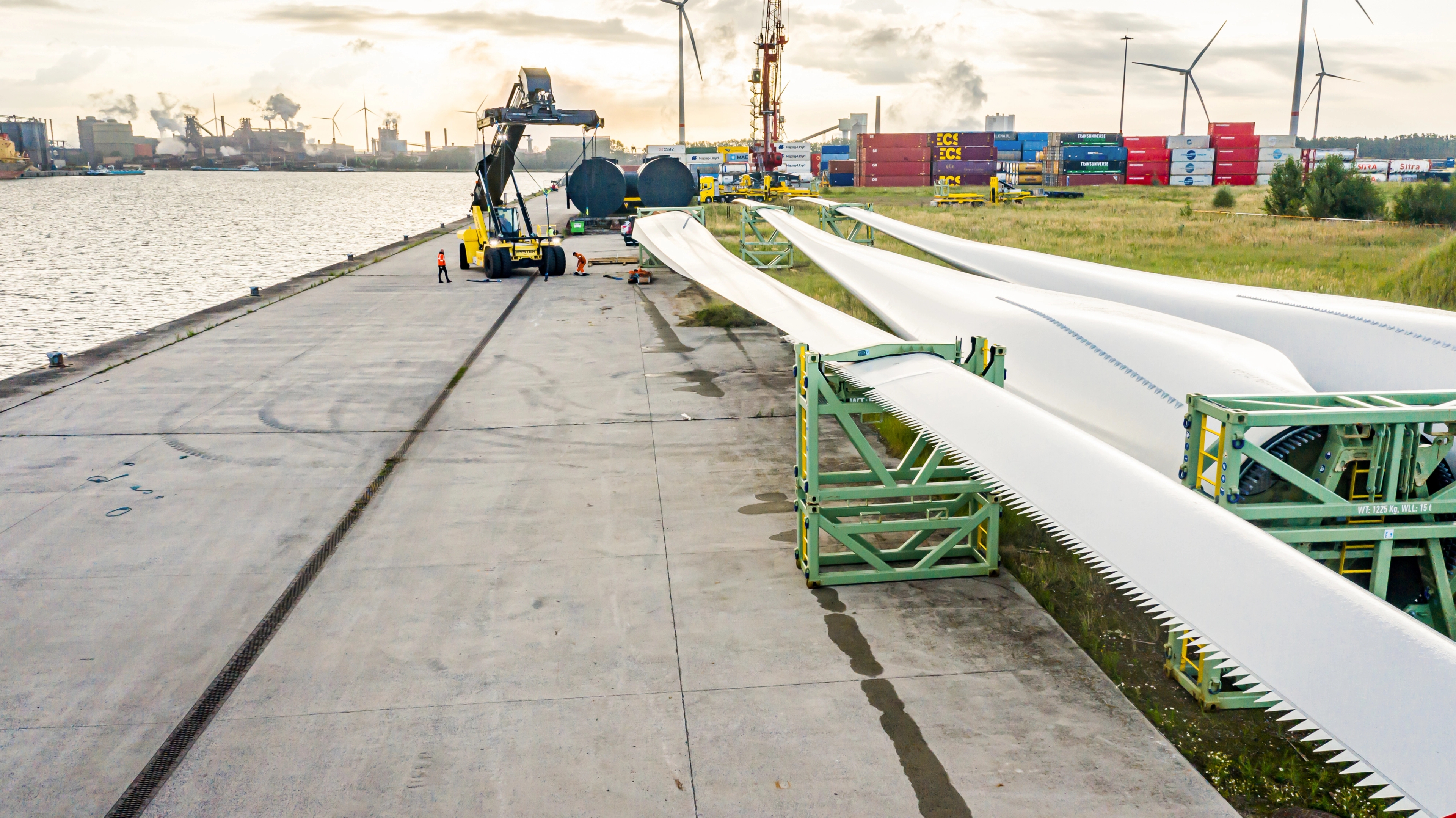 3 pales d'éoliennes sont disposées sur un quai industriel au bord de l'eau