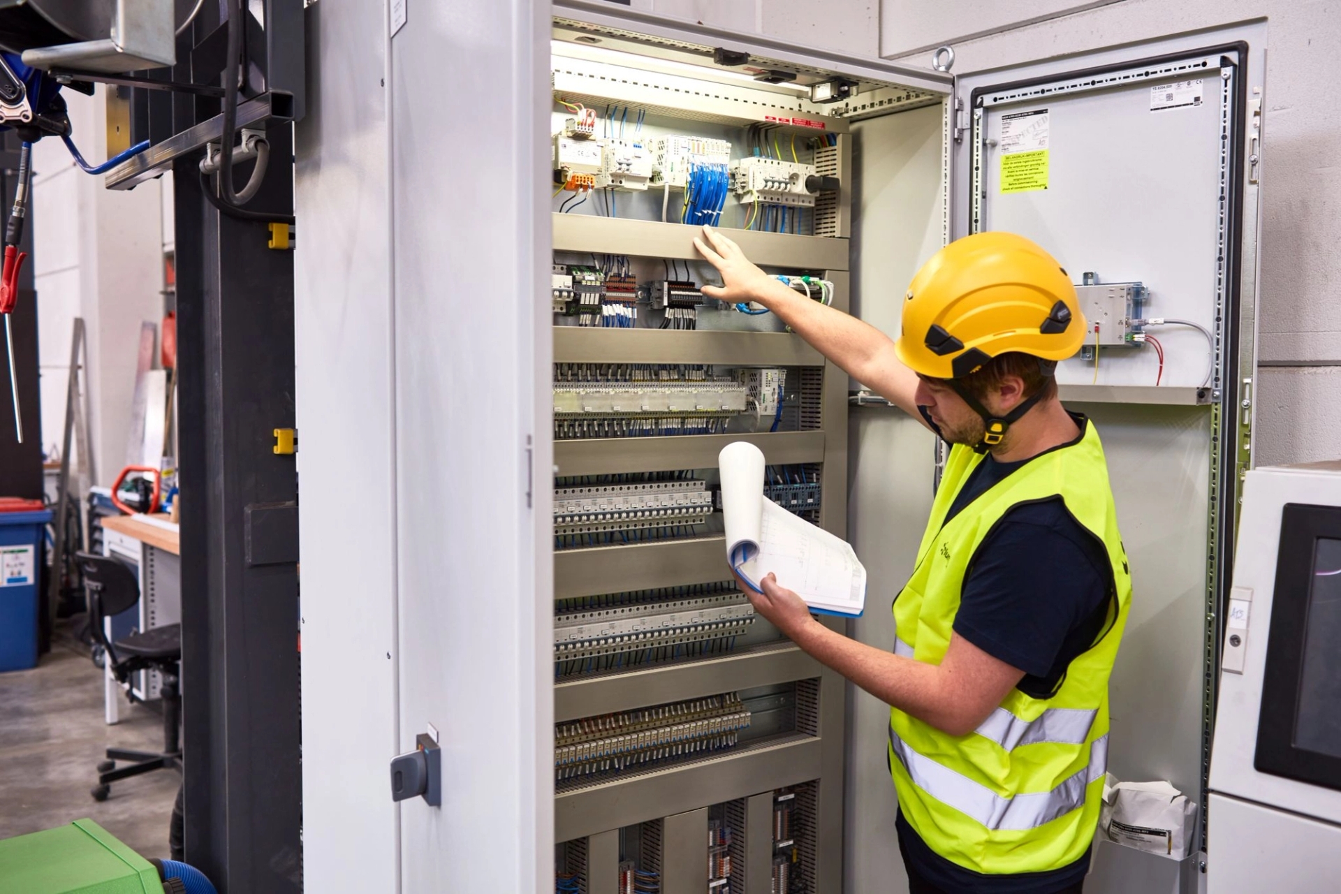 Un technicien en gilet et casque de sécurité inspecte un panneau électrique ouvert dans un environnement industriel