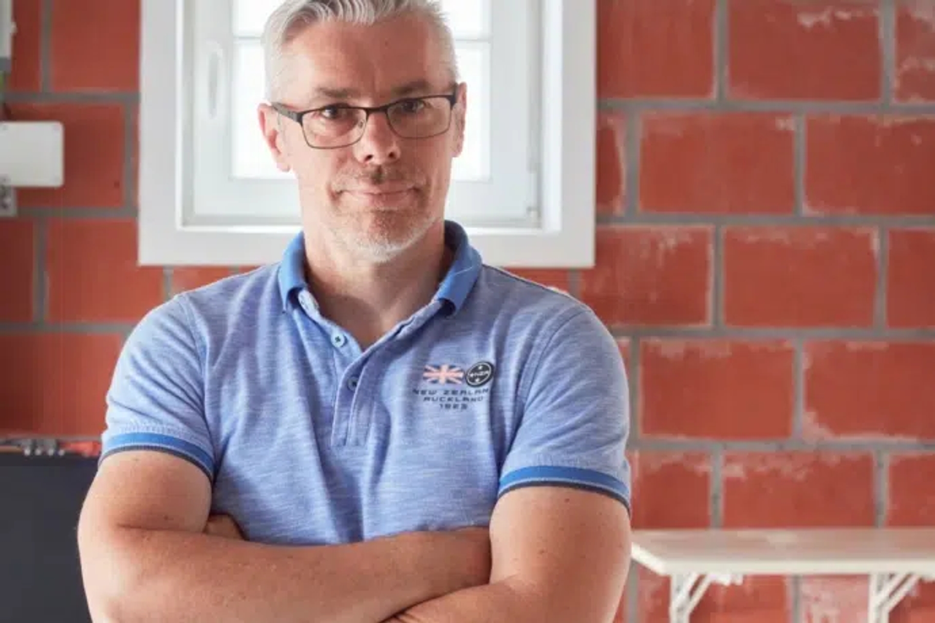 Homme aux cheveux gris, portant un polo bleu clair et des lunettes, dans son garage à côté de sa batterie domestique.