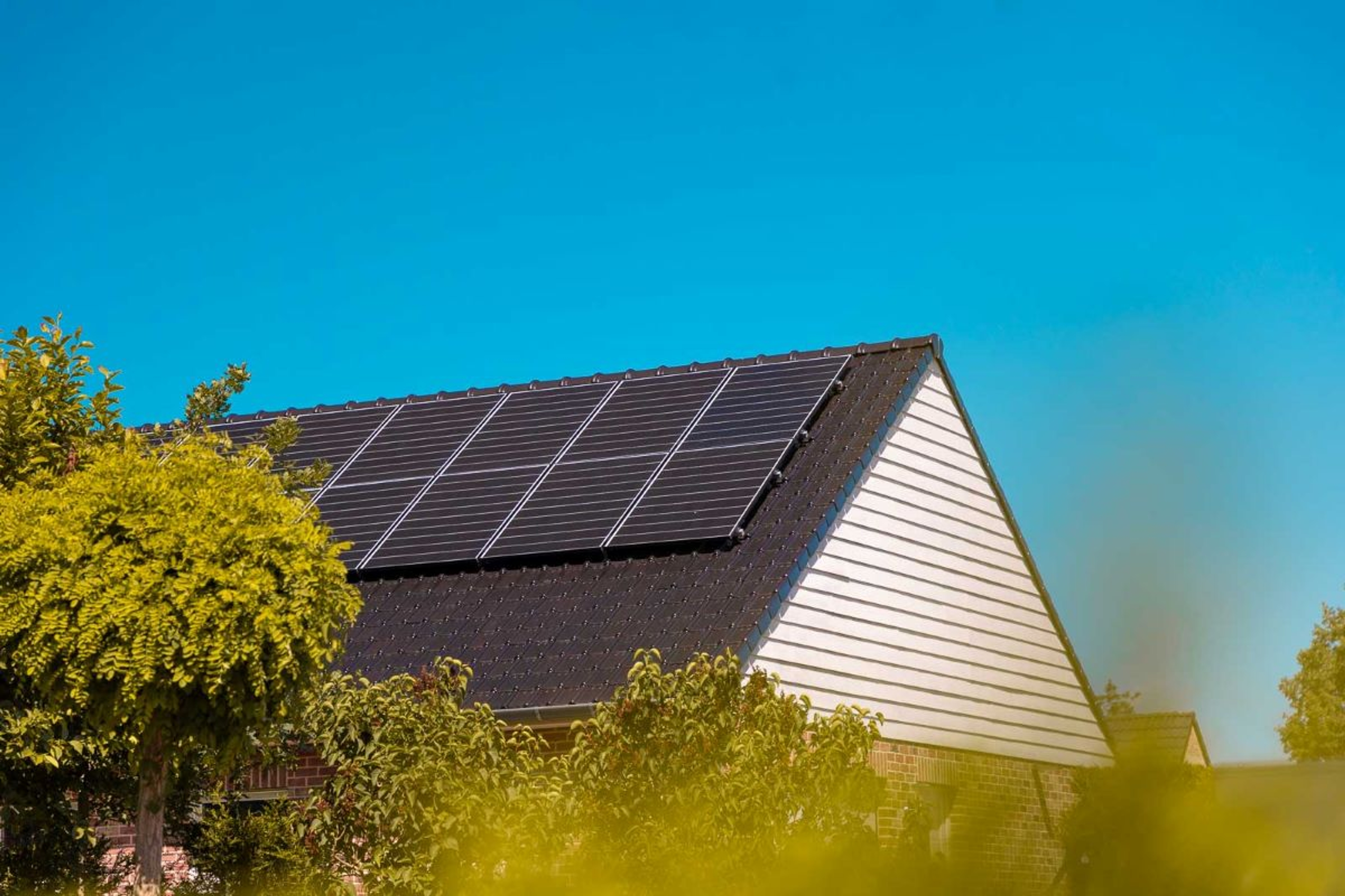 Toit en pente d’une maison avec des panneaux solaires noirs installés, entourée de végétation et sous un ciel bleu clair